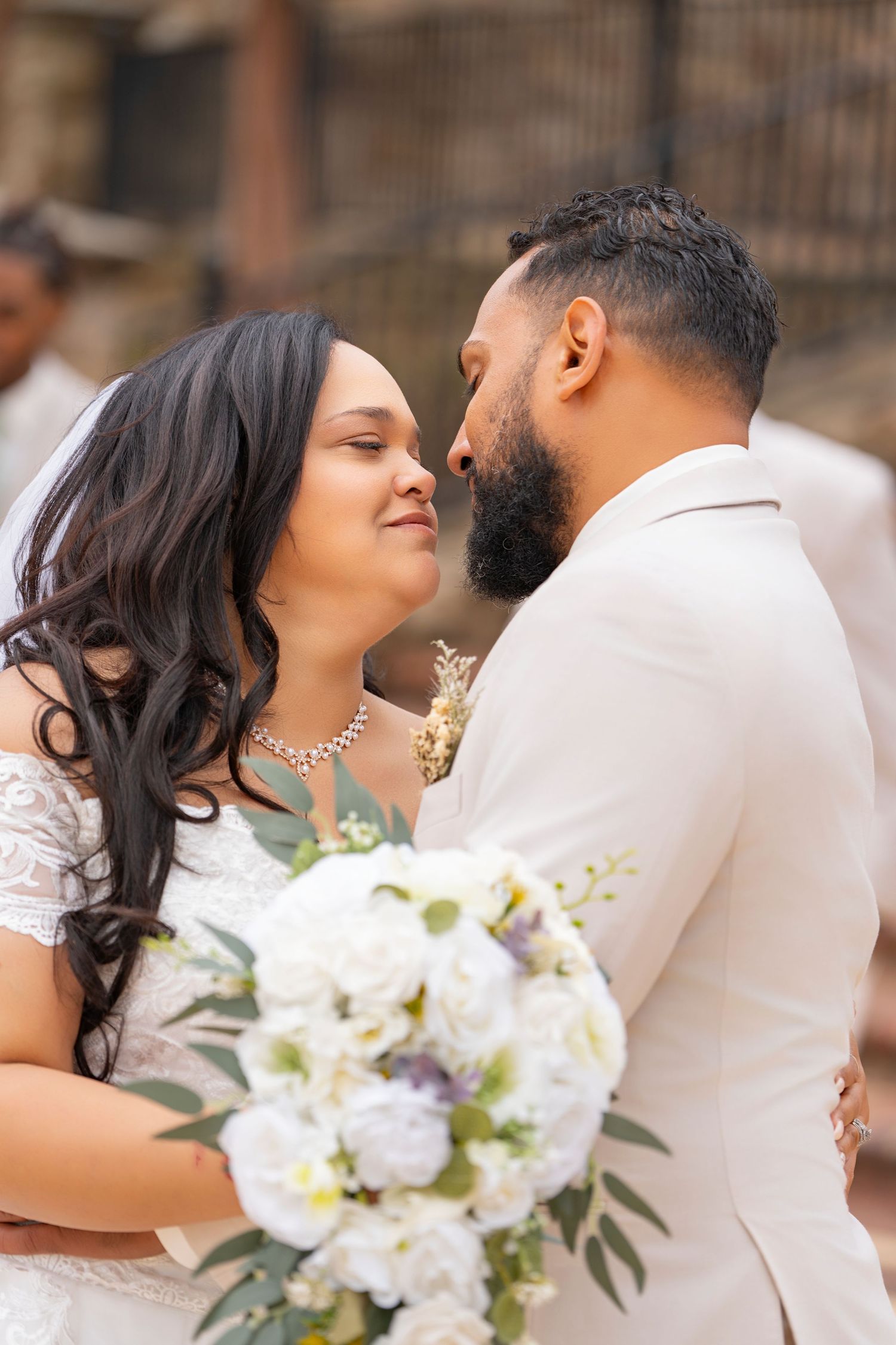 A romantic wedding couple shares tender moments while holding a white floral bouquet in an outdoor setting.