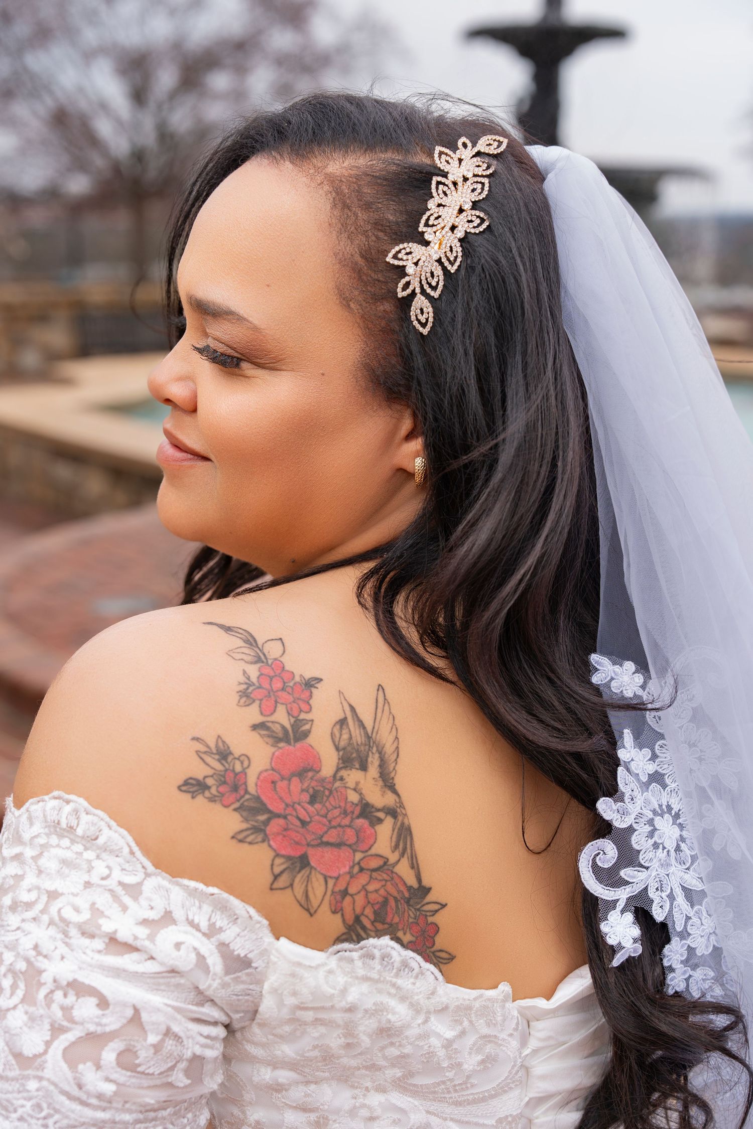 A bride wearing a white lace dress shows off a colorful floral tattoo on her shoulder and back.