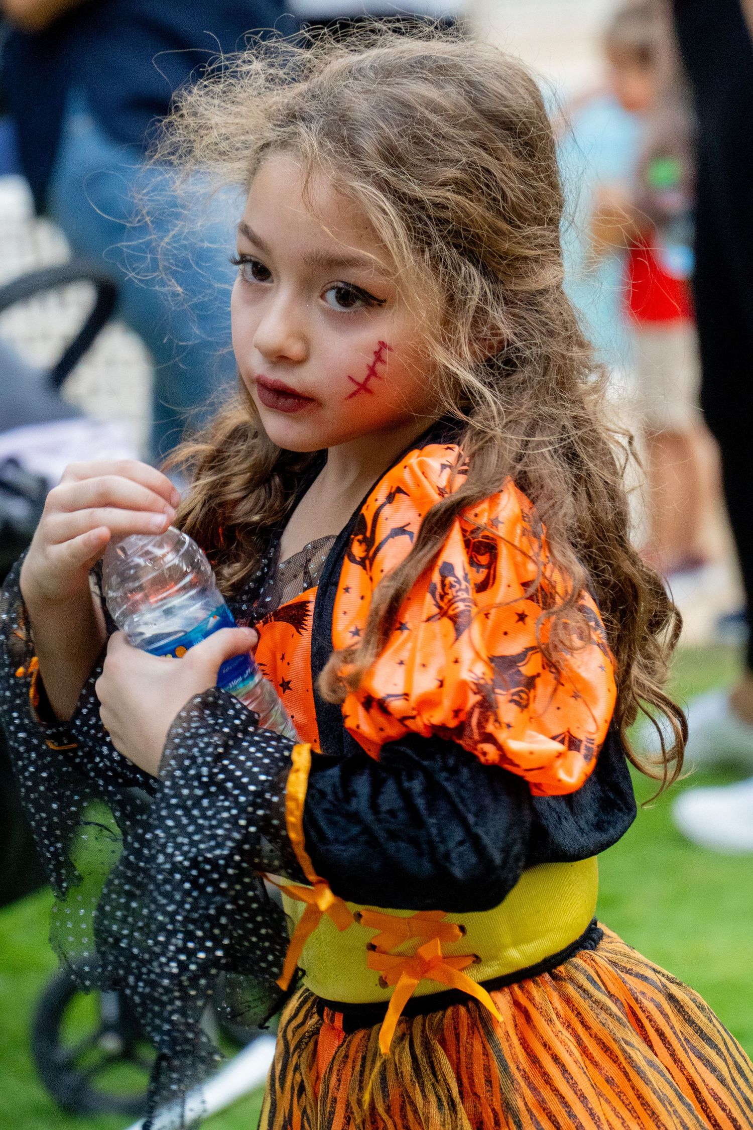 Young person in orange and black Halloween dress with face paint and curly hair.
