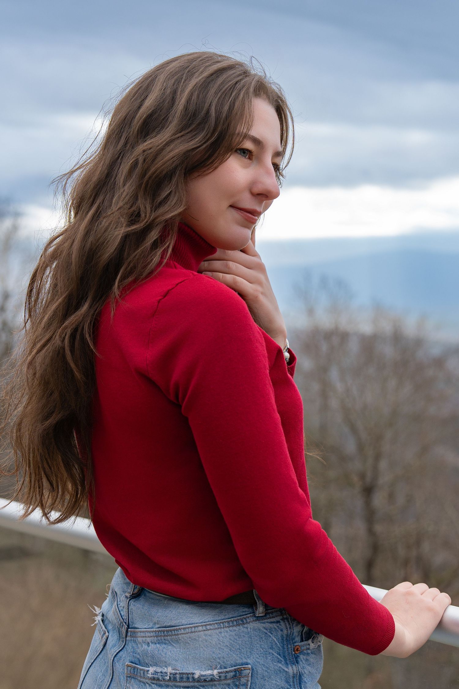 Woman in red sweater and jeans poses thoughtfully against a cloudy winter landscape in a series of outdoor portraits.