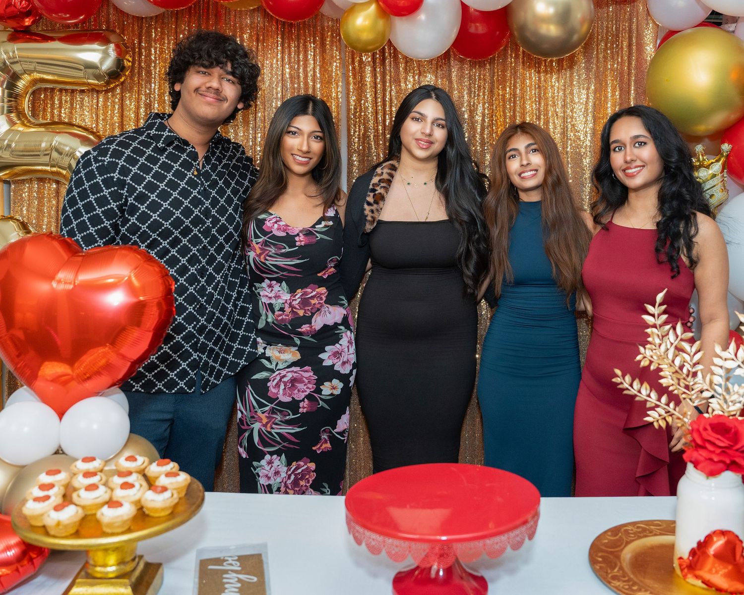 Group photo at a party with gold sequin backdrop, red heart balloon, and dessert table decorations.