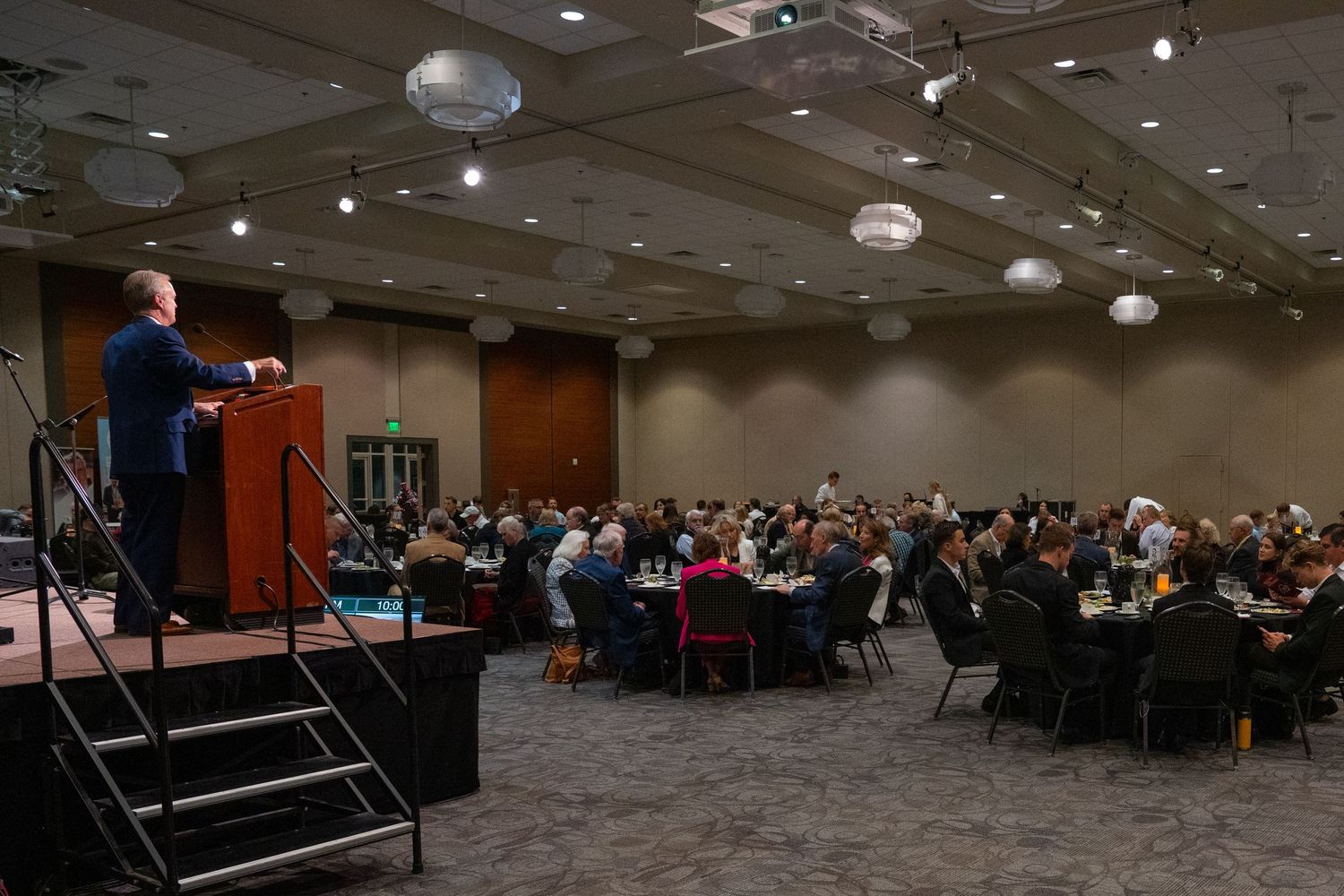 Speaker at podium addresses large audience in conference room with round ceiling lights and theater-style seating.