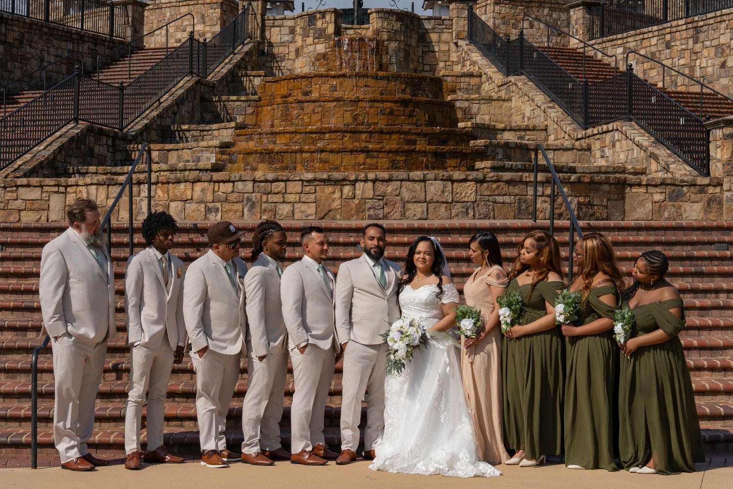 Wedding party stands in formal attire on stone steps with bridesmaids in olive green dresses and groomsmen in gray suits.