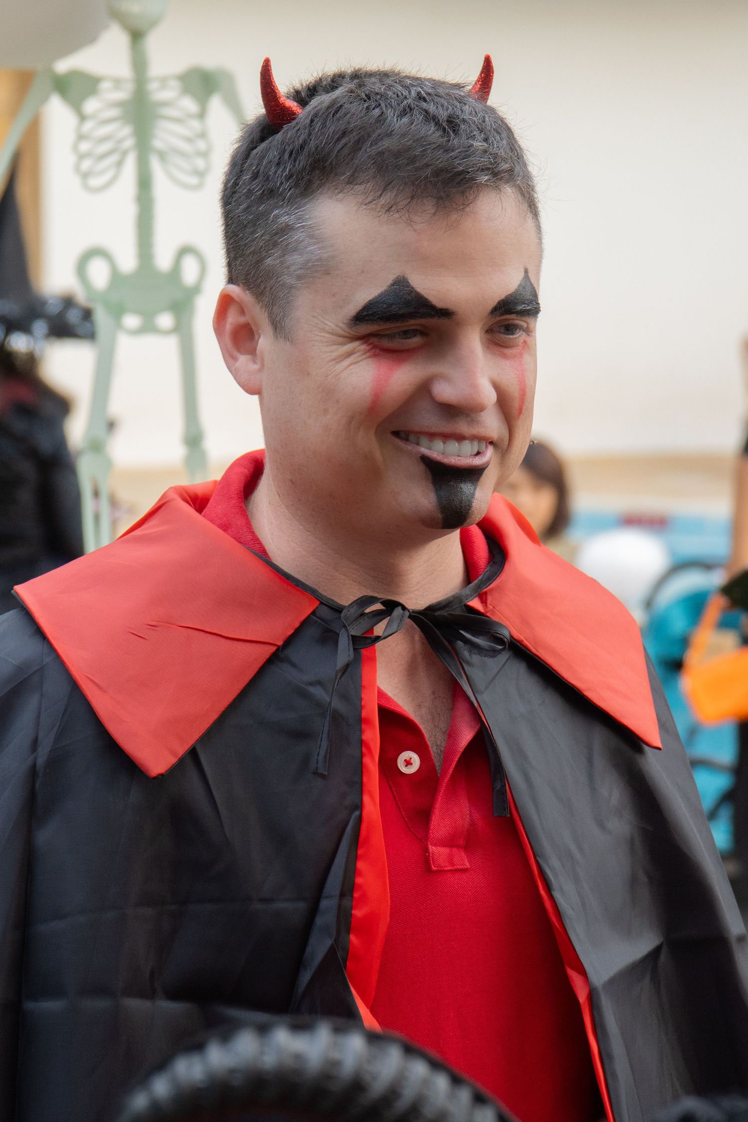 Person in devil costume with red horns and black cape at Halloween party.