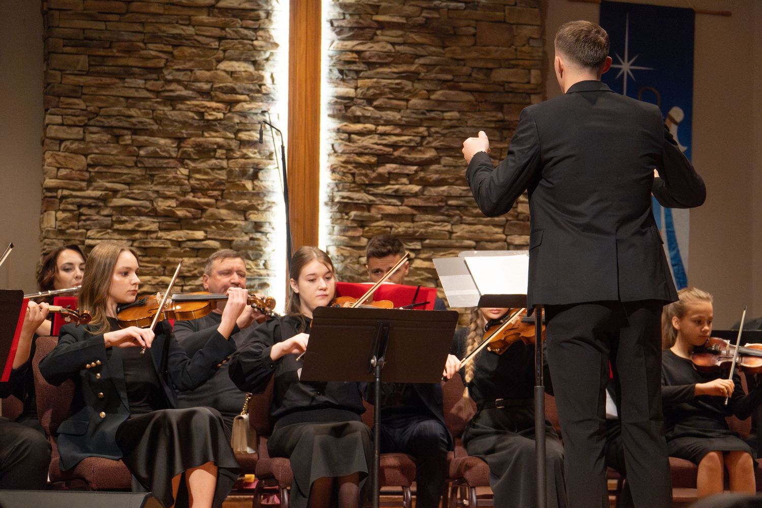 Musicians play string instruments during performance in church with stone wall backdrop.