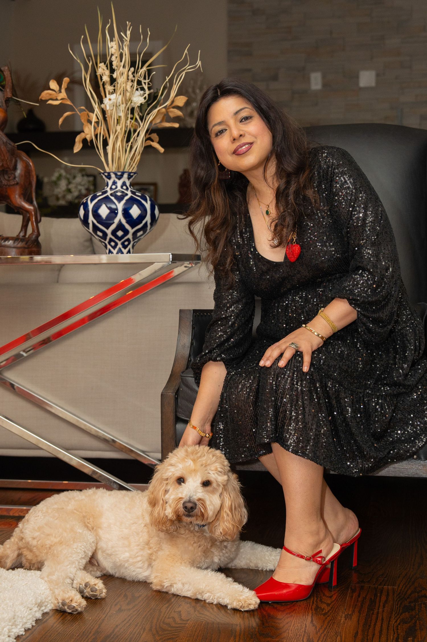 A person in a sparkly black dress poses with a fluffy goldendoodle dog on hardwood floors near red shoes and decor.