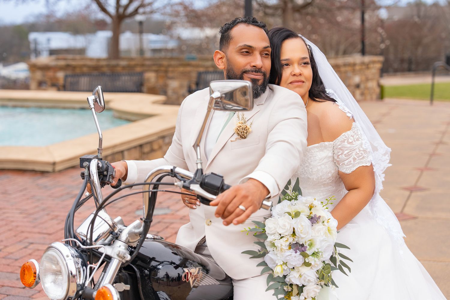 A romantic wedding couple poses with a motorcycle, featuring white floral bouquets and elegant attire.