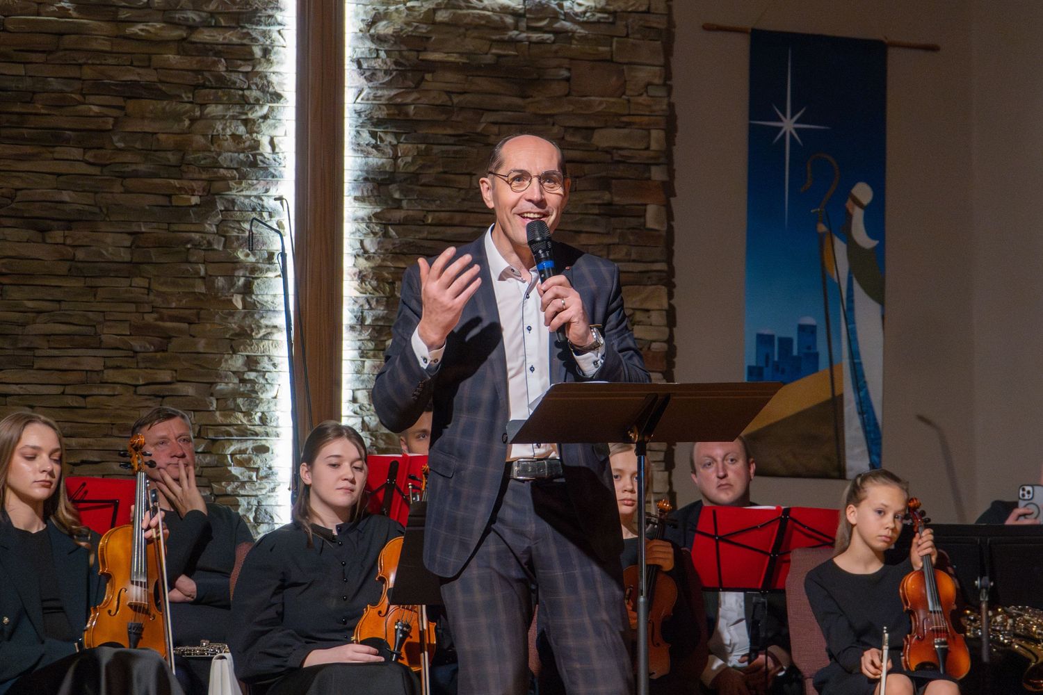 Speaker in grey suit gestures while presenting to orchestra in church setting with stone wall backdrop.