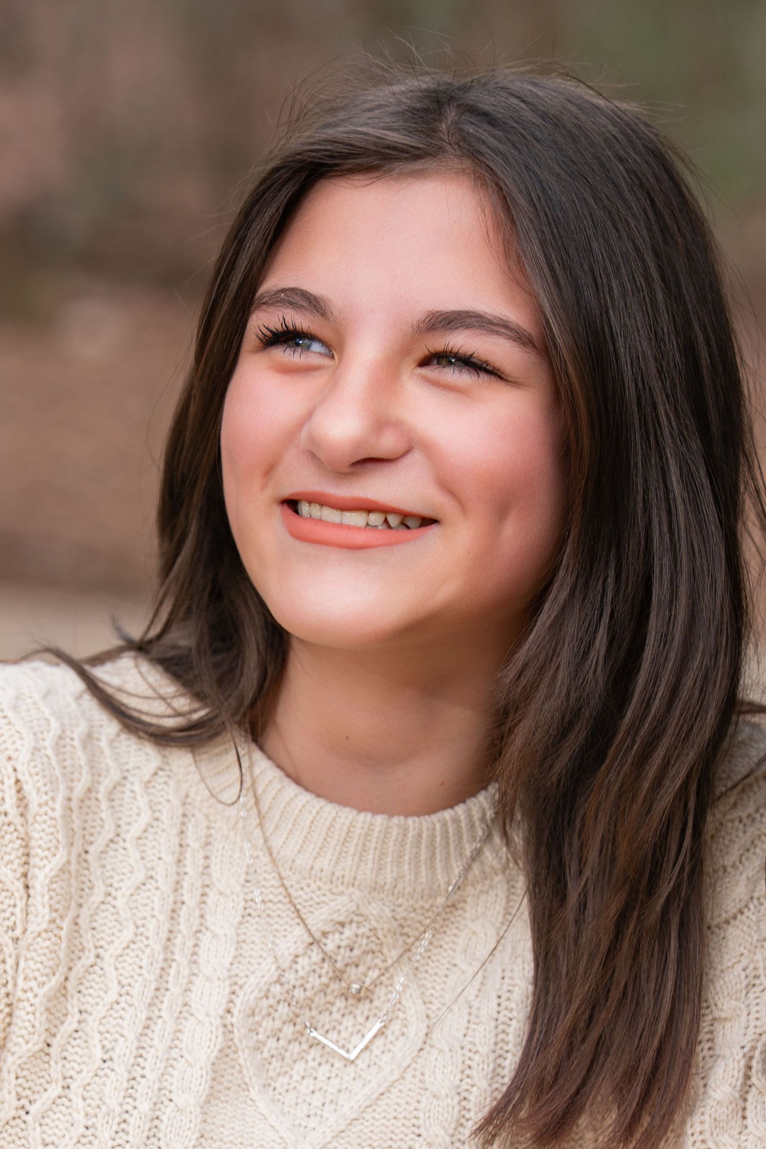 A person wearing a cream knit sweater smiles warmly while posing outdoors against a natural blurred background.