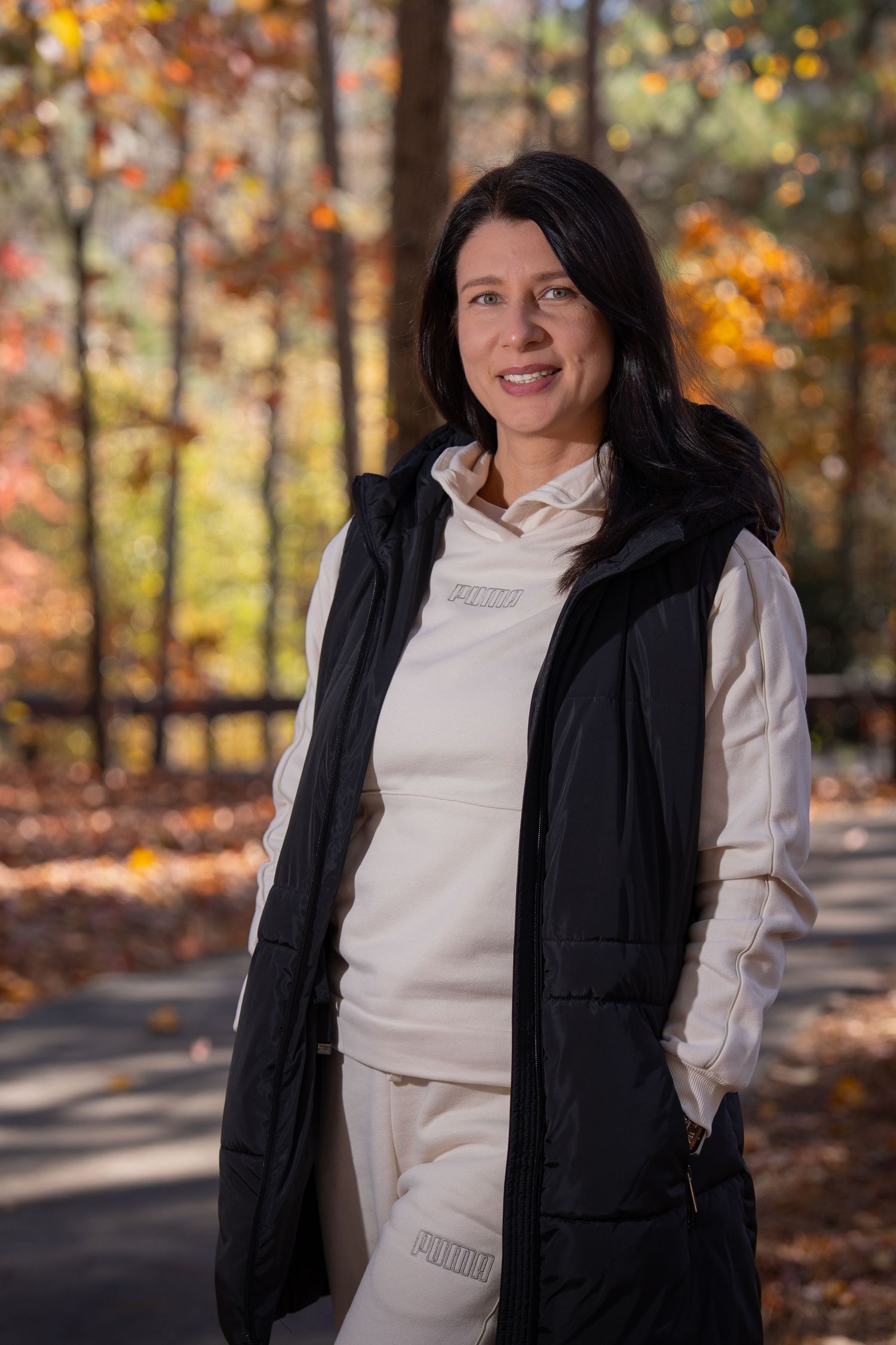 Person wearing white athleisure and black vest poses on autumn path with fall foliage.