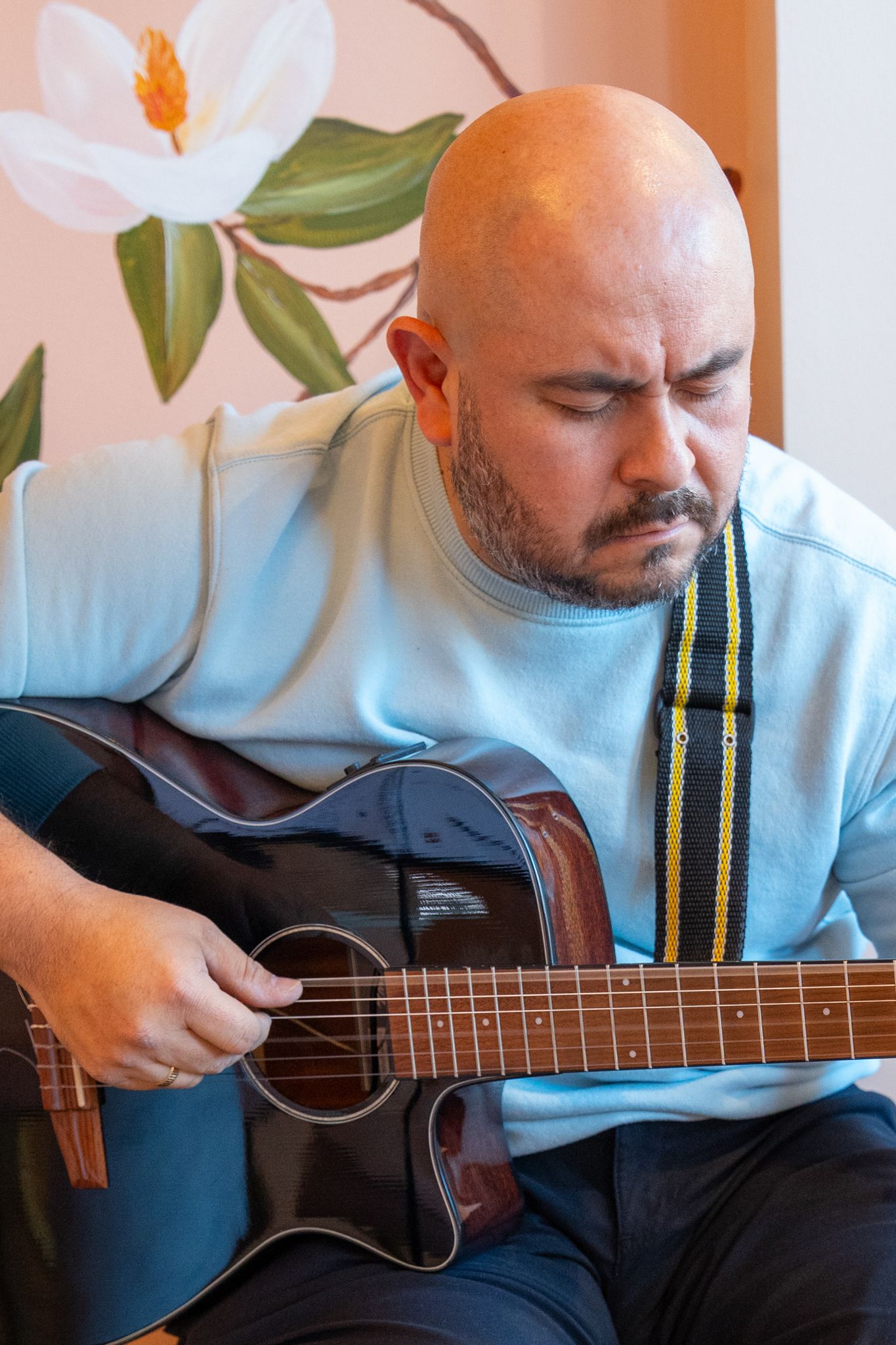 Person in light blue shirt playing acoustic guitar against floral wallpaper background.