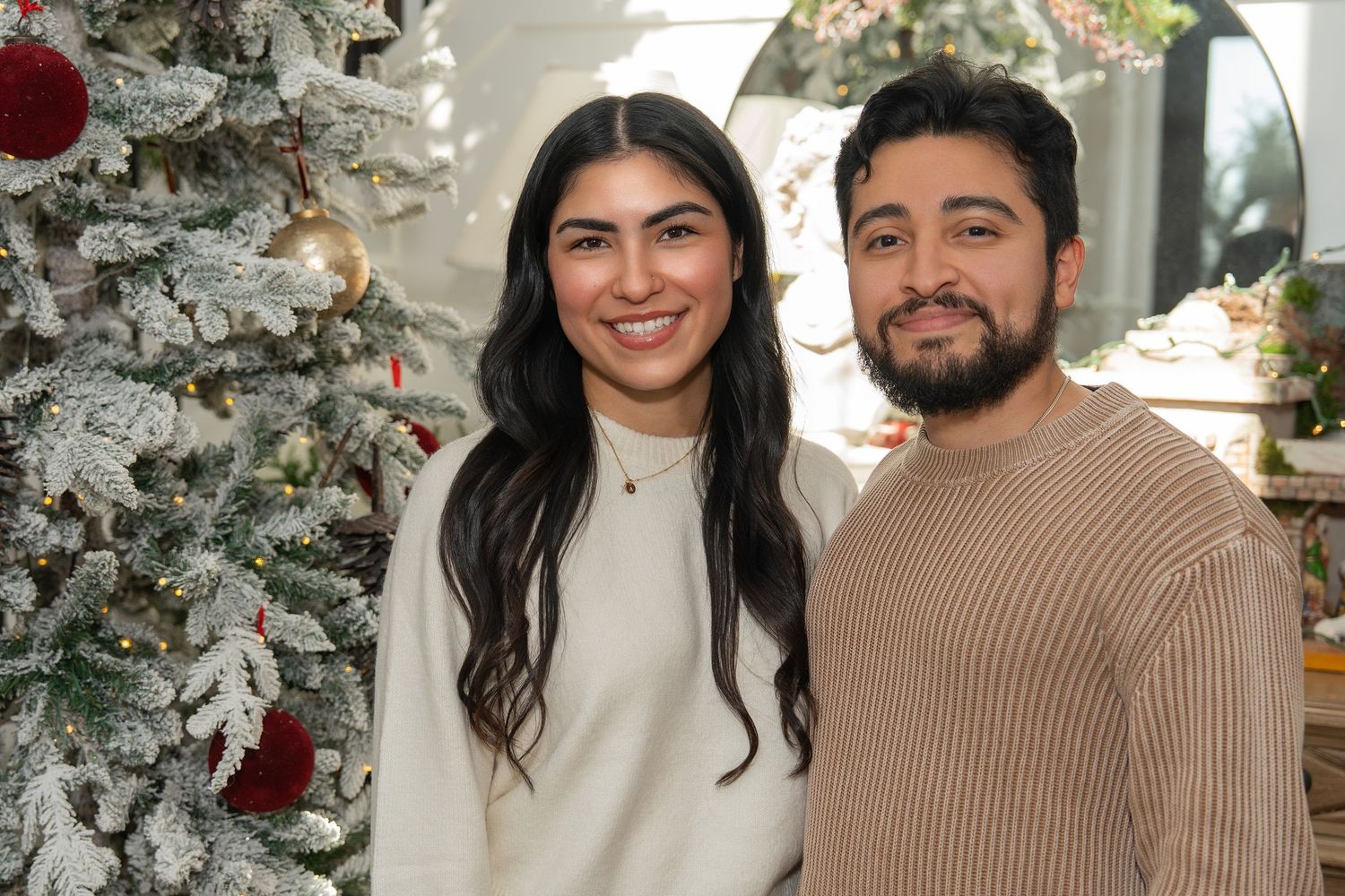 A couple poses together in front of a decorated Christmas tree with white and silver ornaments.