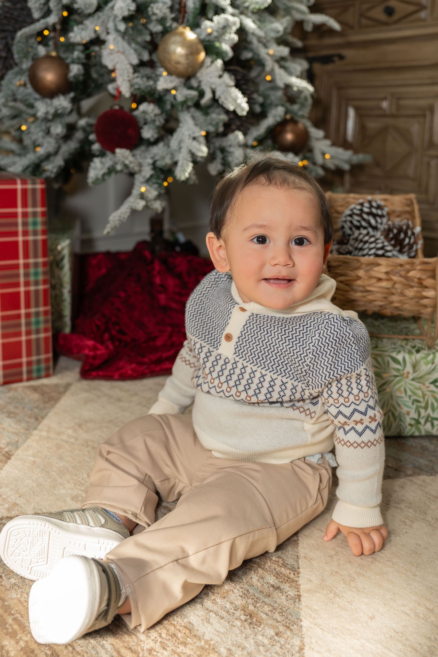 A baby in a beige outfit and patterned scarf lies on a white rug near a decorated Christmas tree.
