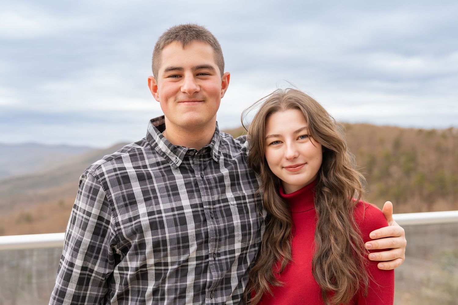 A couple in plaid and red sweater poses together outdoors against a scenic mountain landscape backdrop.