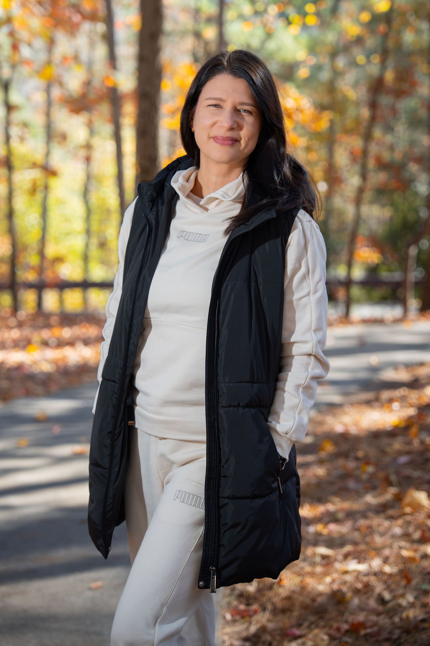 Person in white loungewear and long black vest poses on autumn walking trail.