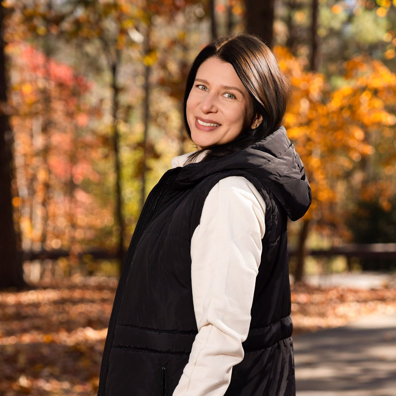 Person wearing a black vest and white sweater poses in front of vibrant autumn foliage on a forest path.