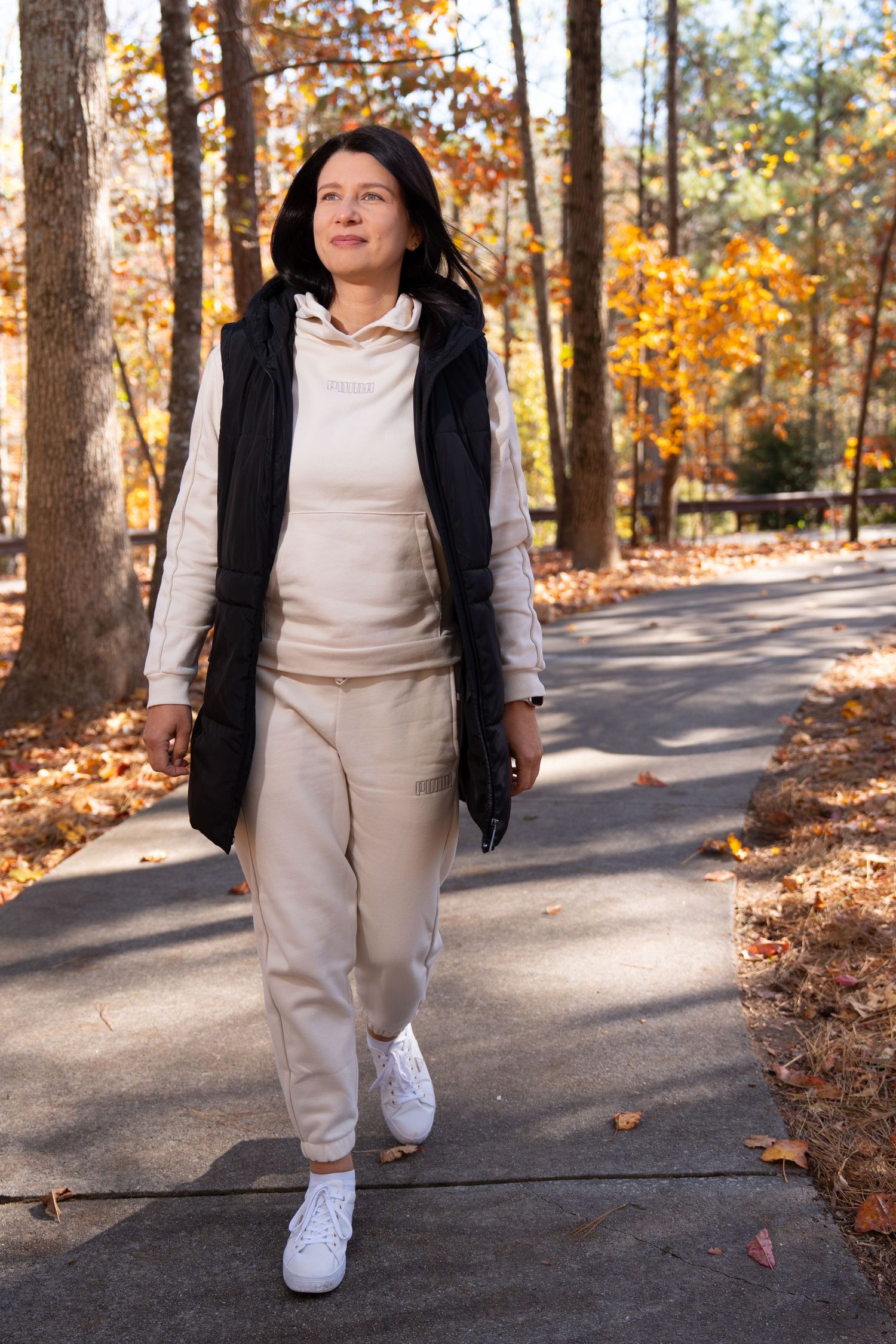 Person in white outfit and black vest walking on autumn trail through wooded area.