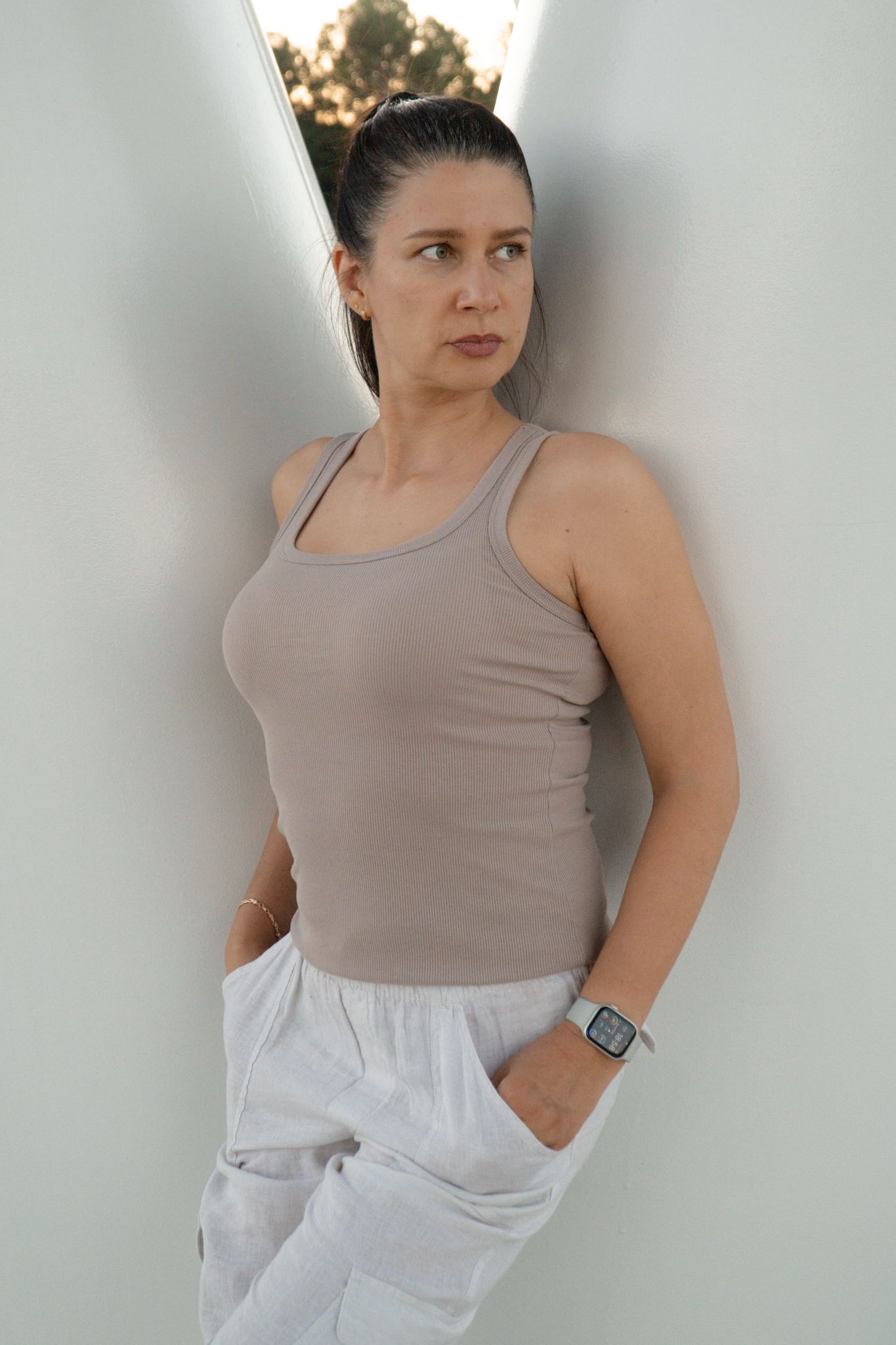 Person wearing beige tank top and white pants standing against light background.