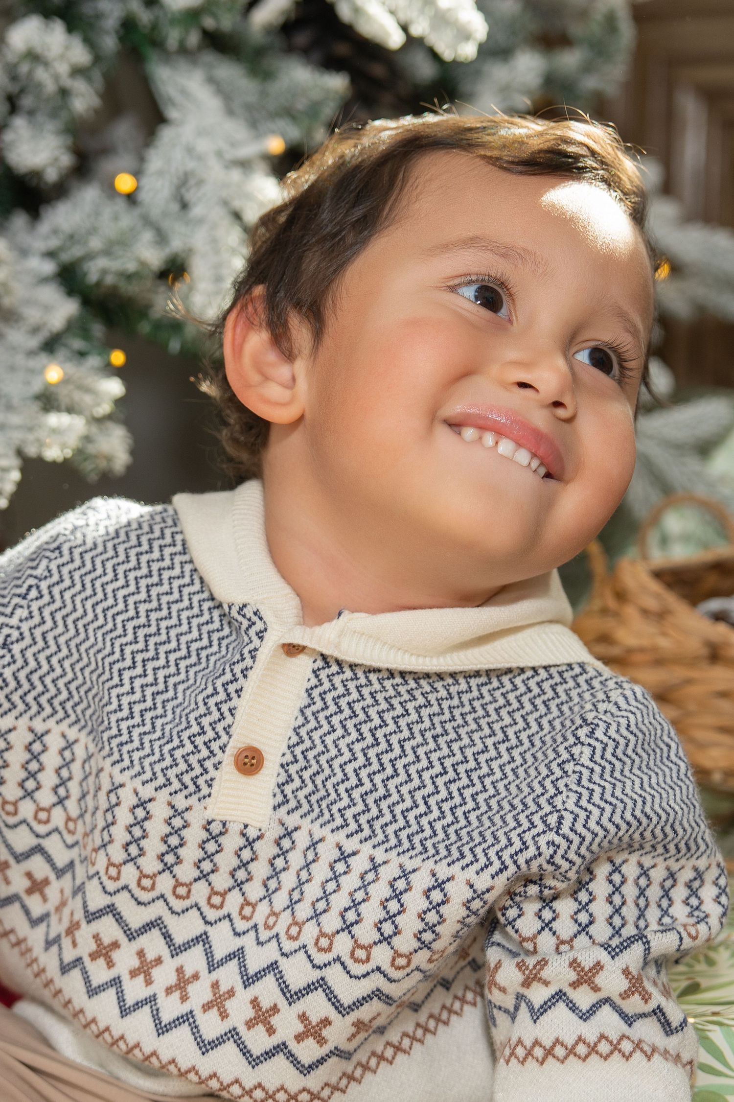 A young child wearing a cozy winter sweater with zigzag patterns smiles warmly against a festive holiday background.
