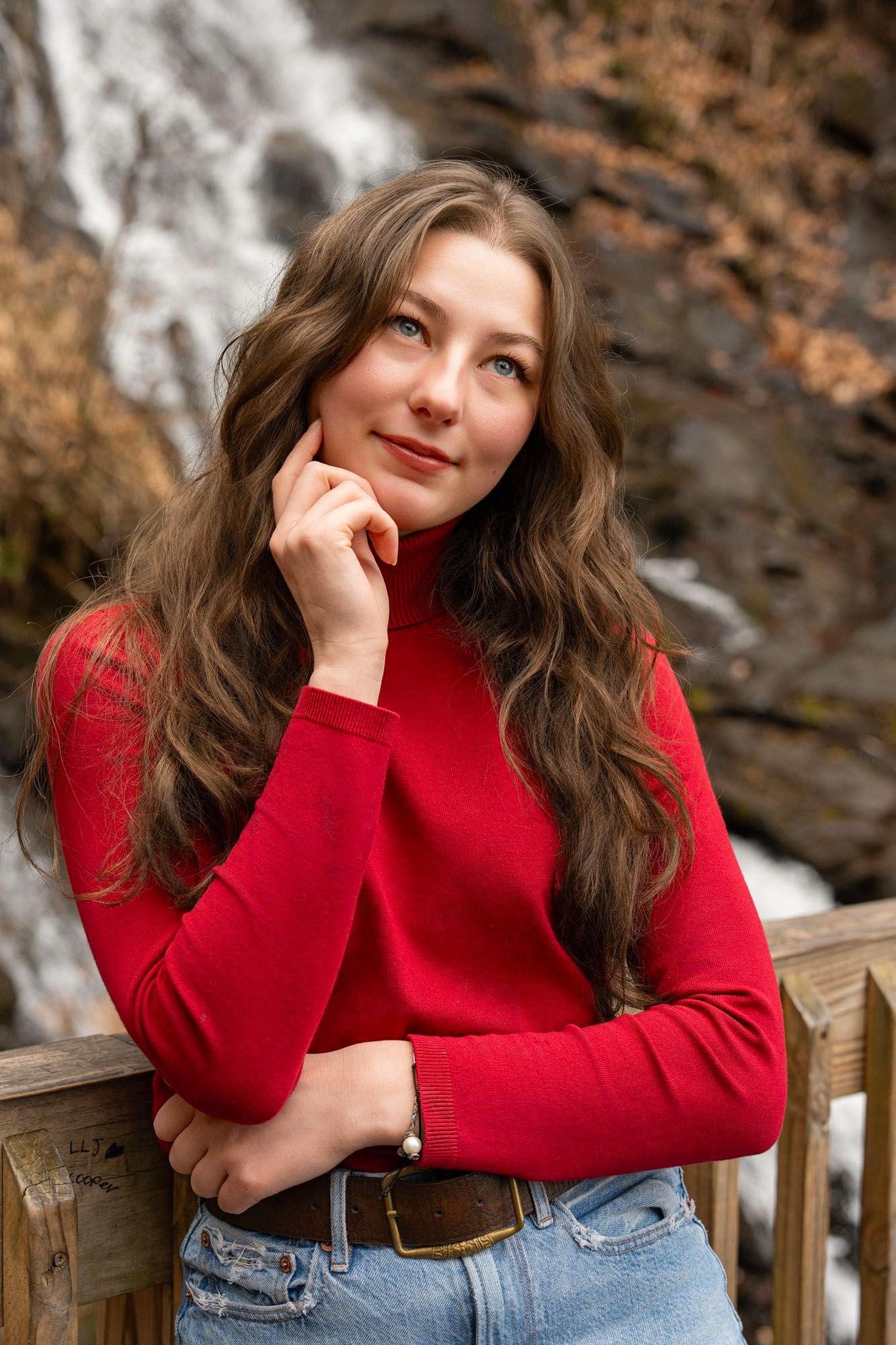 A person in a bright red sweater and jeans poses thoughtfully on a wooden deck with autumn foliage in the background.