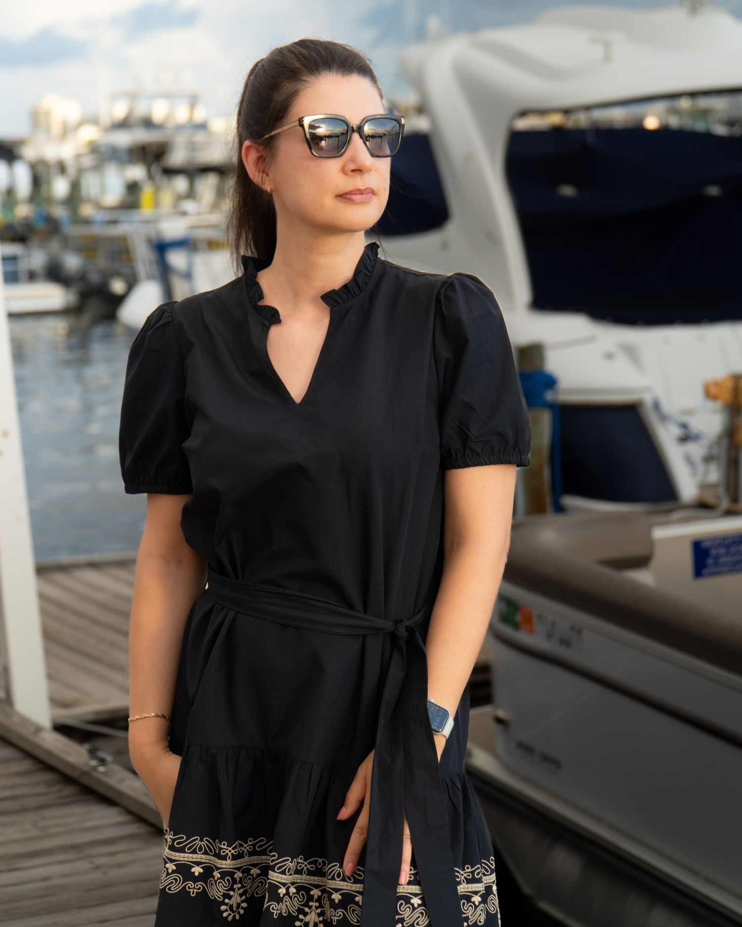 Woman in black dress stands on dock with boats and marina in background.