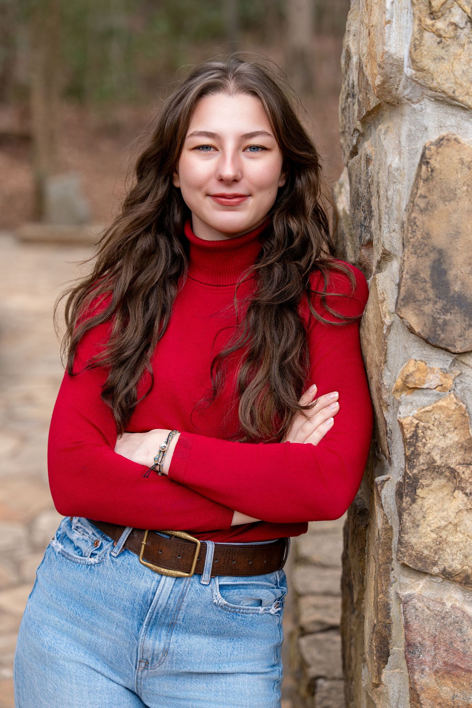 A person in a red turtleneck sweater and blue jeans poses against a stone wall in a natural outdoor setting.