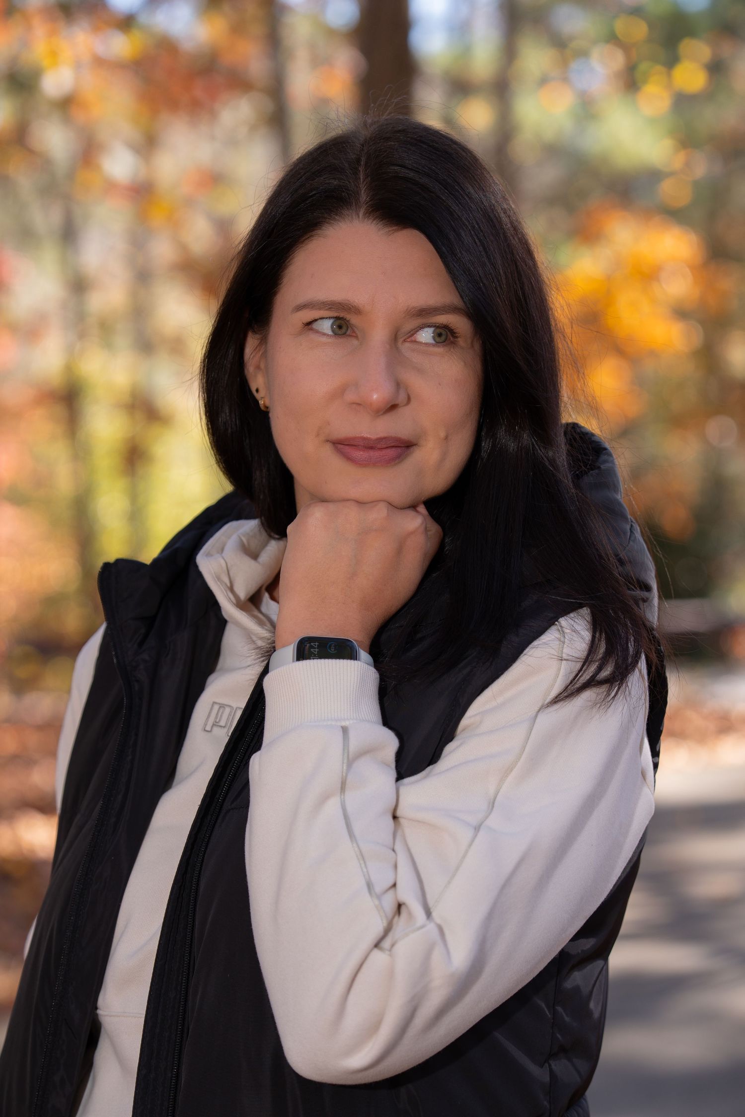 Person in white sweater and black vest poses thoughtfully in autumn setting.