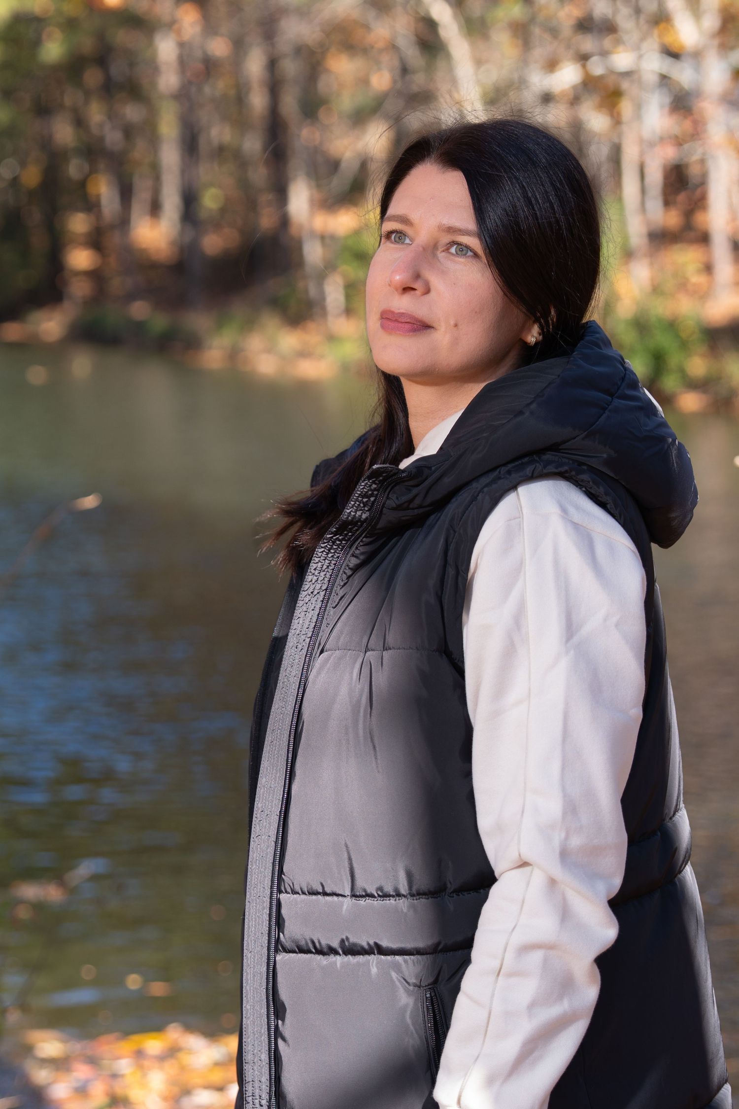 Person wearing black puffy vest and white top poses against autumn lake backdrop.