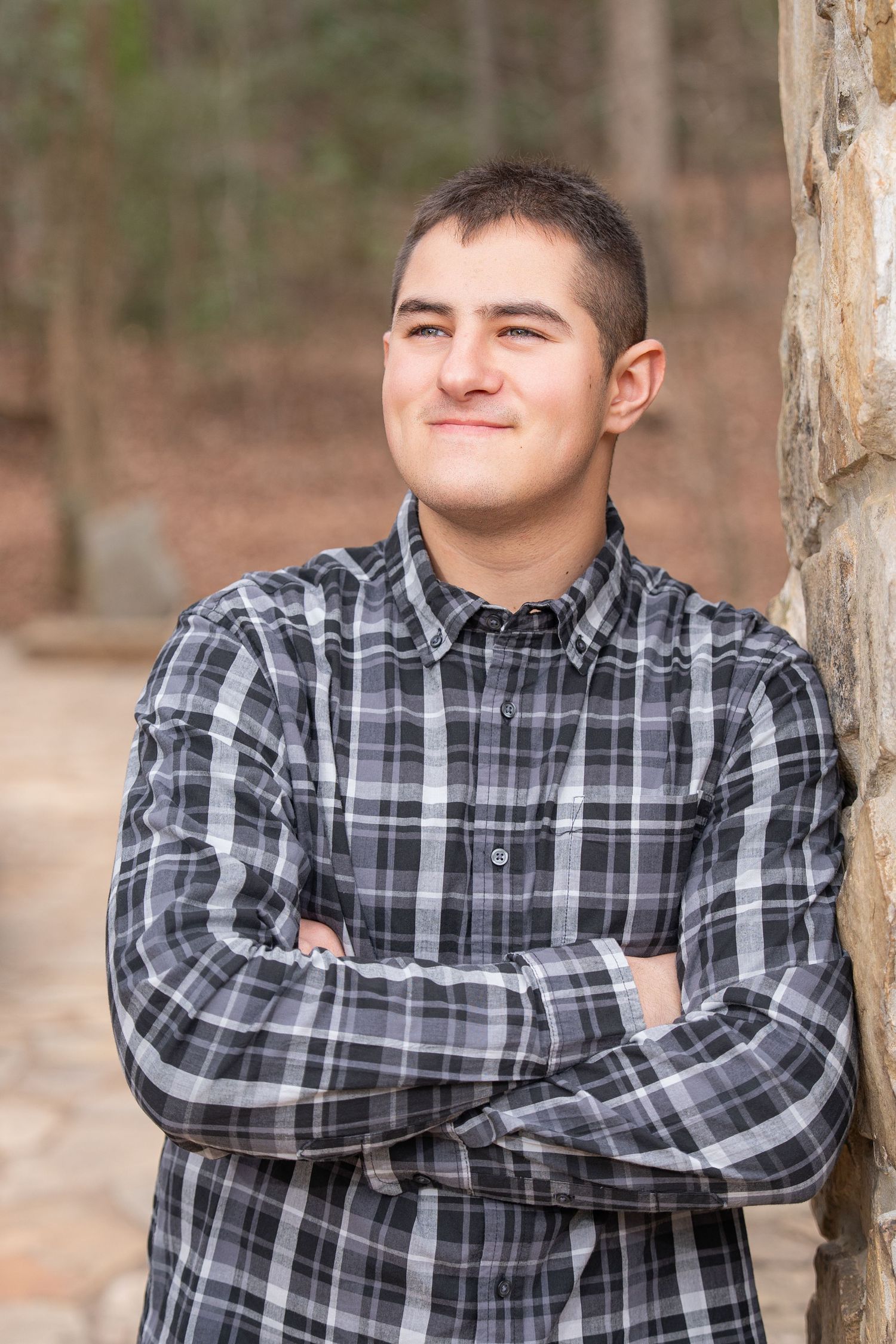 Person wearing a plaid flannel shirt leans against a tree trunk in an outdoor setting with a neutral expression.