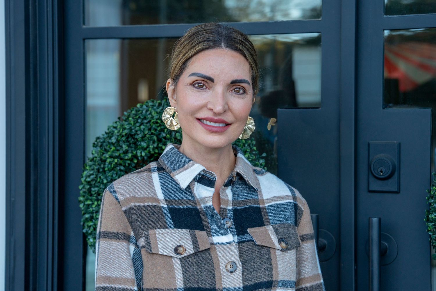 A person wearing a plaid shacket and statement earrings stands in front of a black door with decorative greenery.