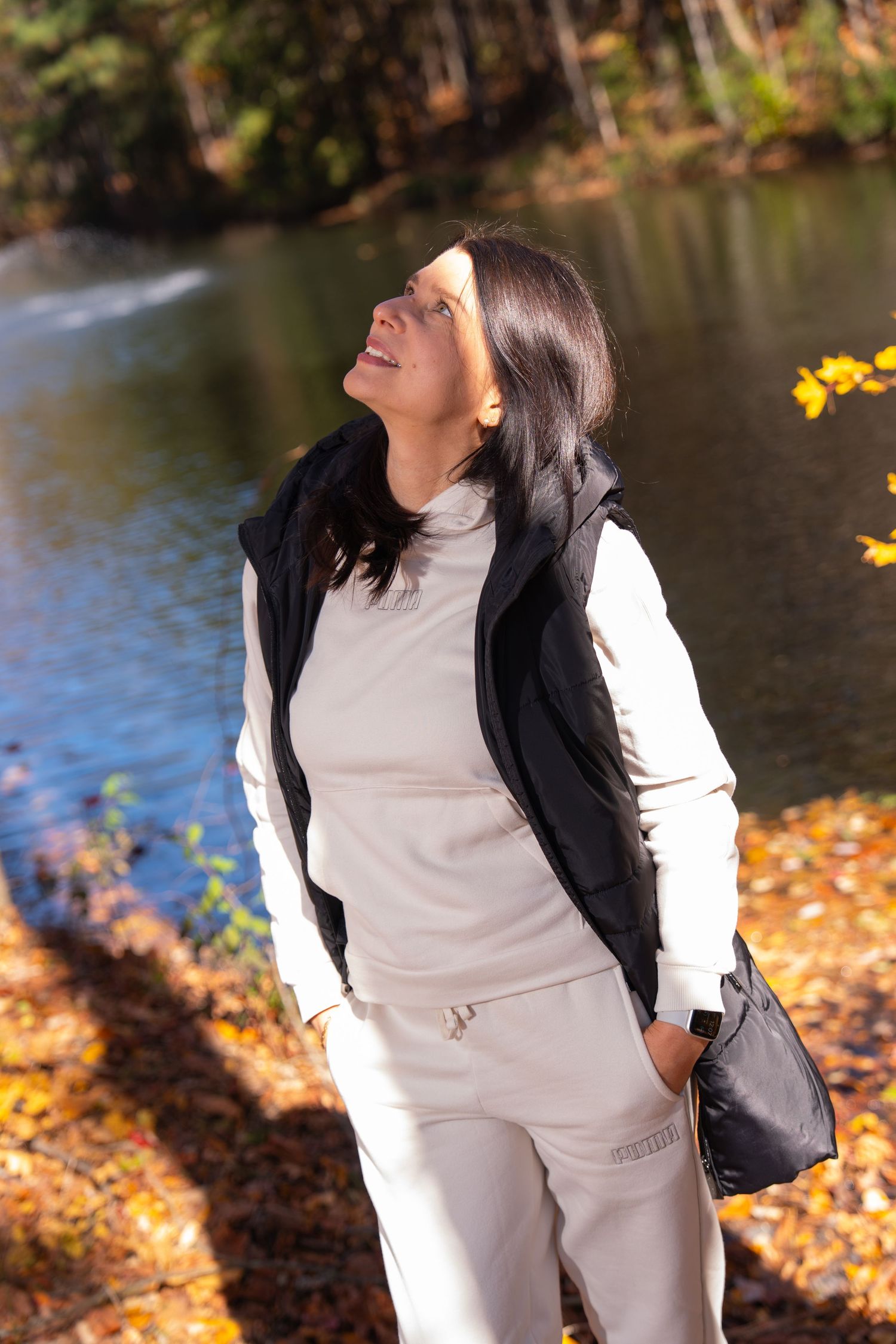 Person in casual white outfit and black vest enjoying fall scenery by a lake.