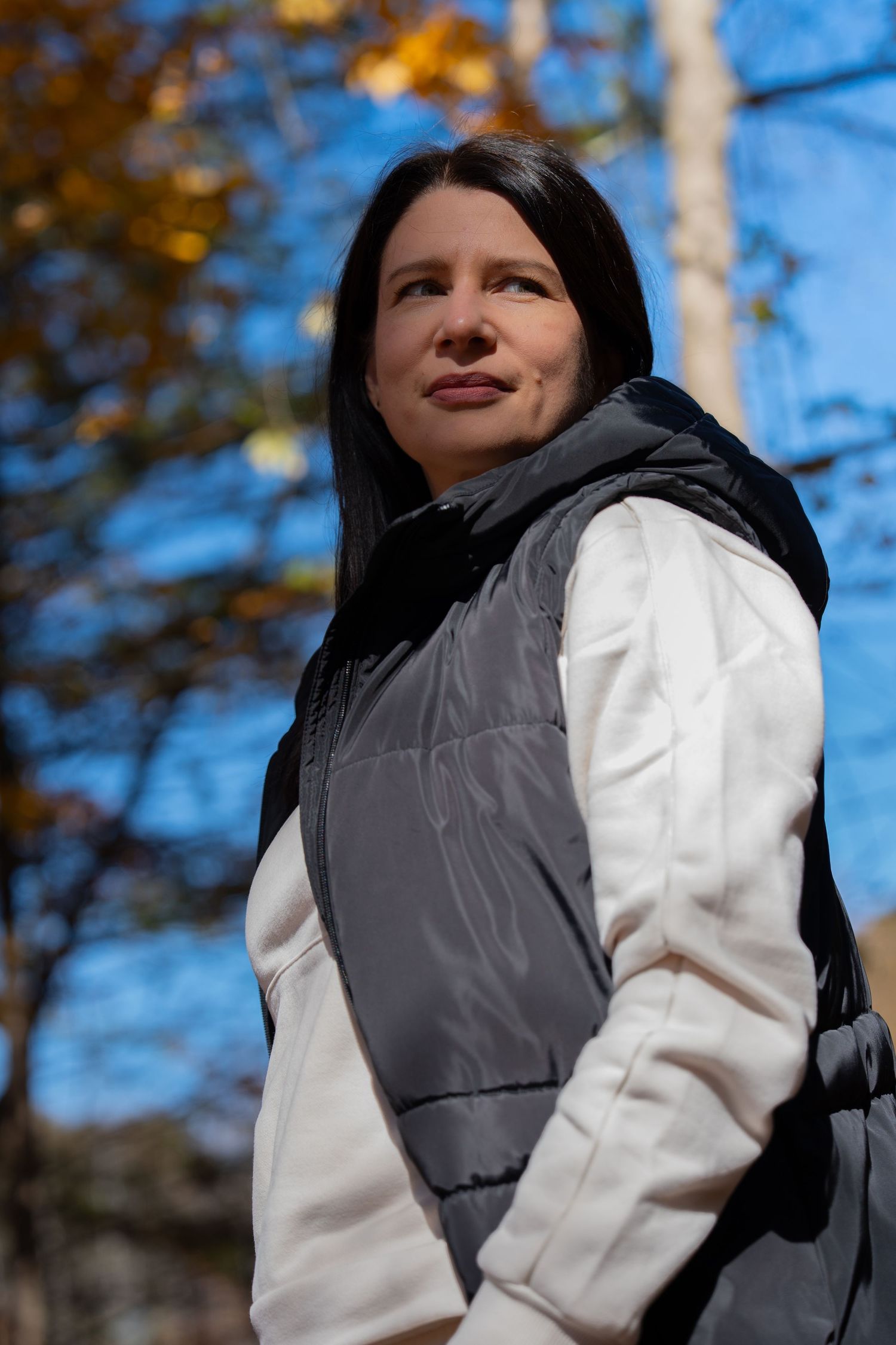 Person in monochrome outfit looking up at blue sky framed by autumn trees.