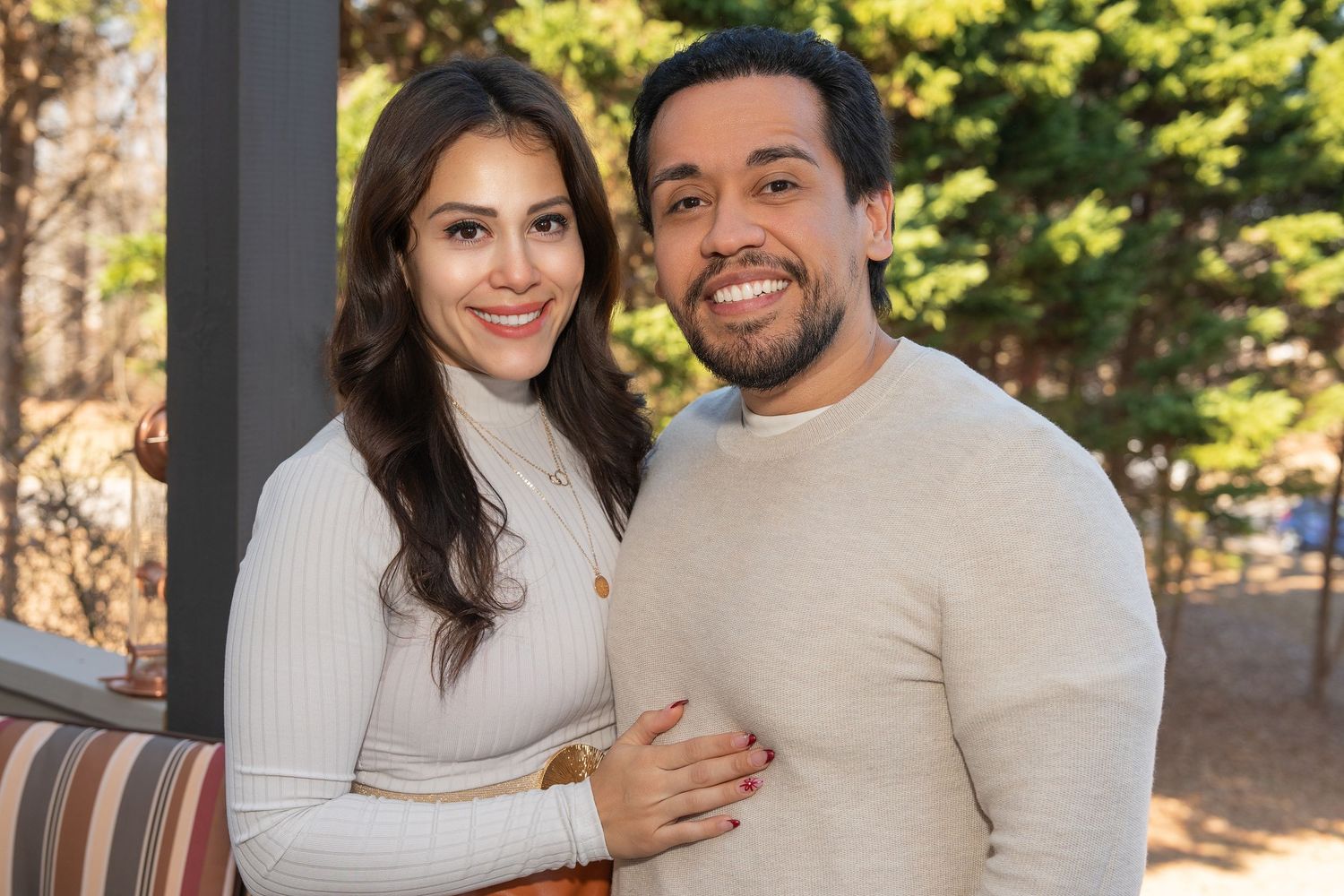 A couple wearing cream colored sweaters embraces outdoors in a natural setting with sunlit trees in the background.