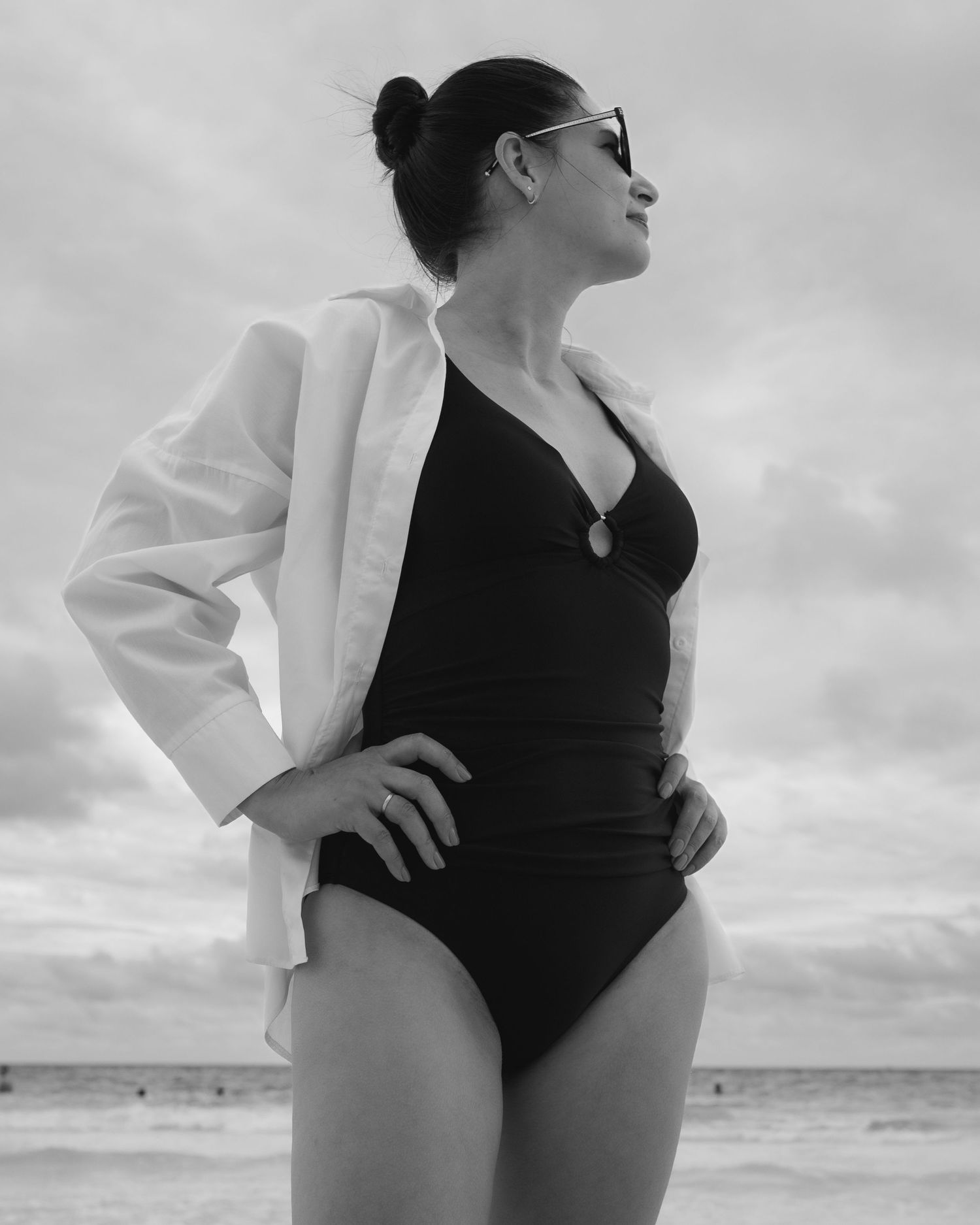 Woman poses in classic black swimsuit and white cover-up against cloudy beach backdrop.