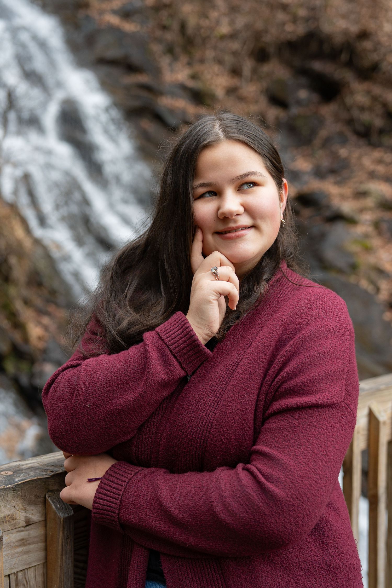 A person in a burgundy sweater poses outdoors against a waterfall backdrop in a casual portrait setting.