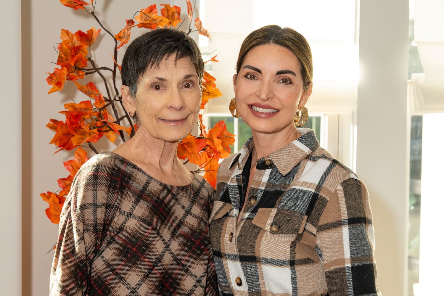 Two people pose together with autumn decorations and orange leaves in a bright interior setting.