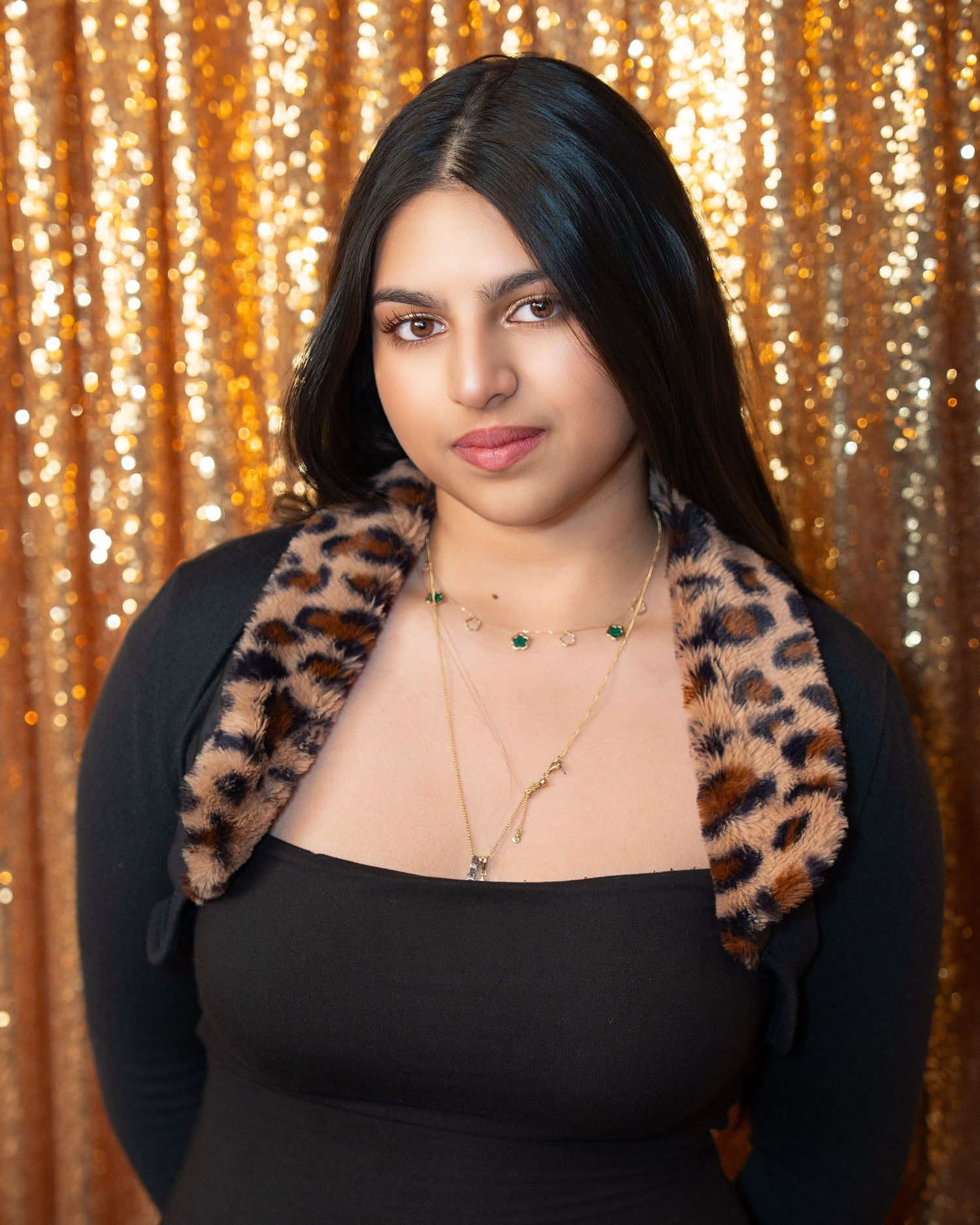 Woman in black dress with leopard print trim poses against gold sequin backdrop.