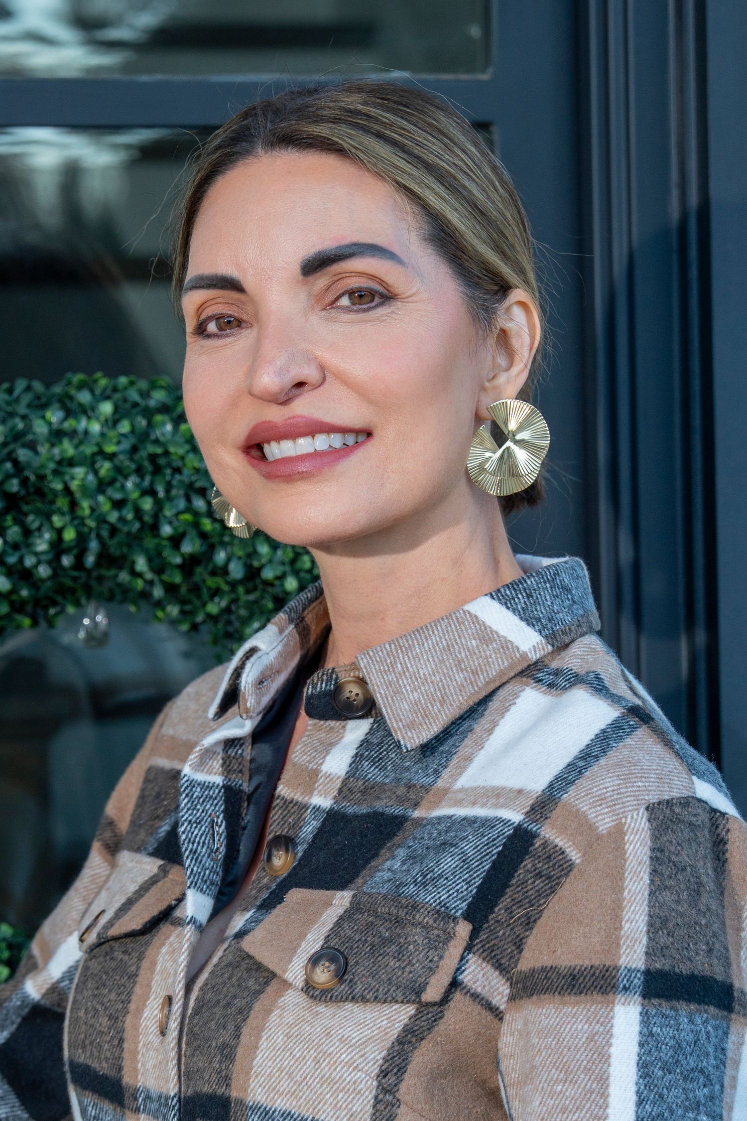 A person wearing a cozy plaid shearling jacket smiles warmly while posing outdoors against a dark background.