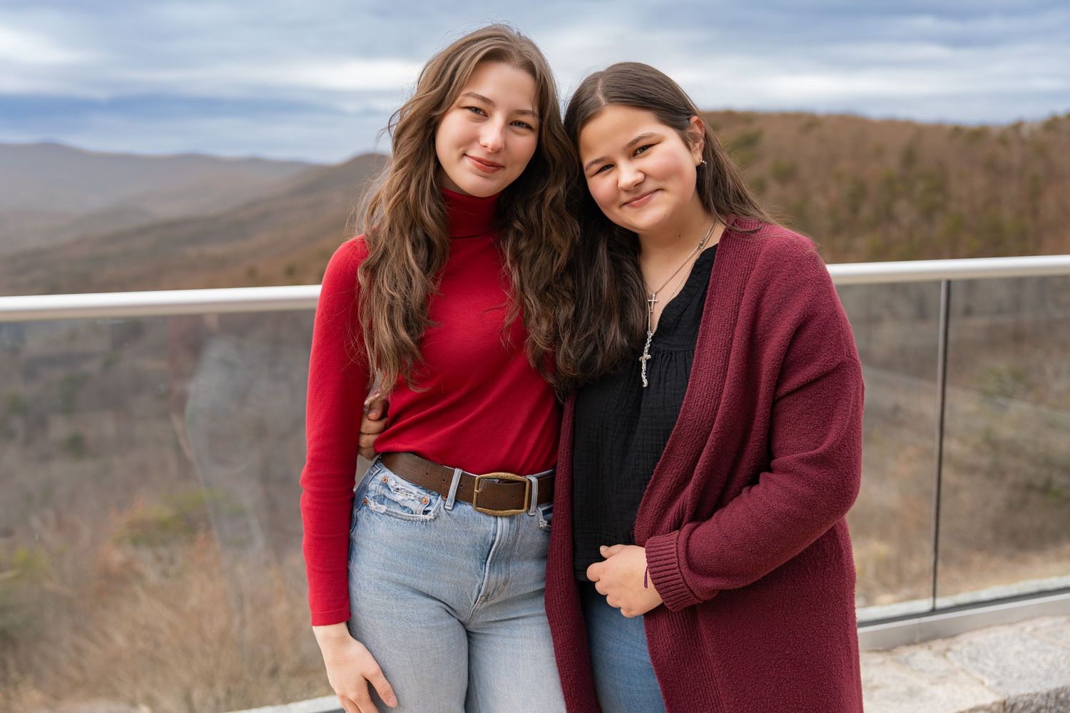 Two people in casual wear pose together against a mountain landscape backdrop.