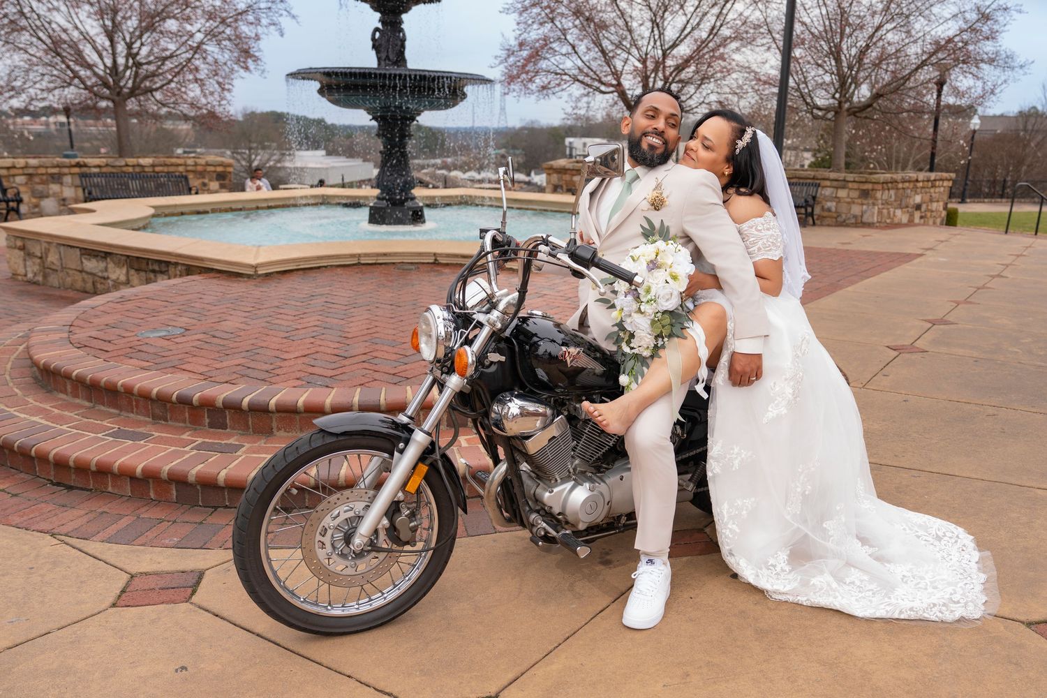 A vintage motorcycle serves as a prop for a romantic outdoor wedding photoshoot in a park setting.