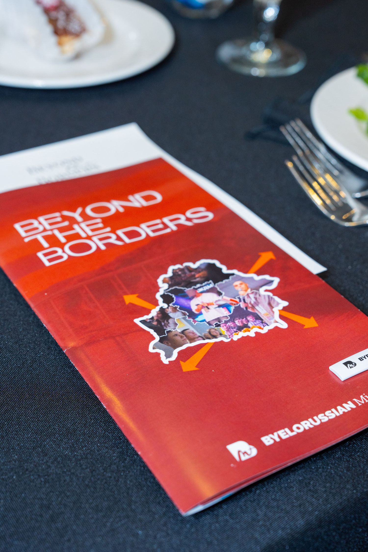 A red program booklet titled 'Beyond the Borders' with a colorful logo sits on a dark table with dining settings.