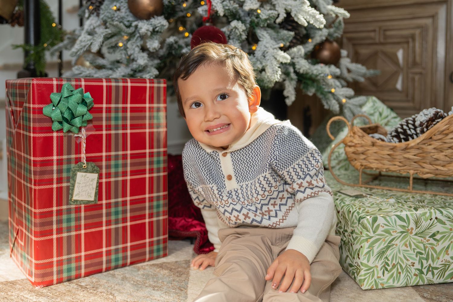 A child in a gray sweater sits by a decorated Christmas tree with wrapped presents in plaid paper.