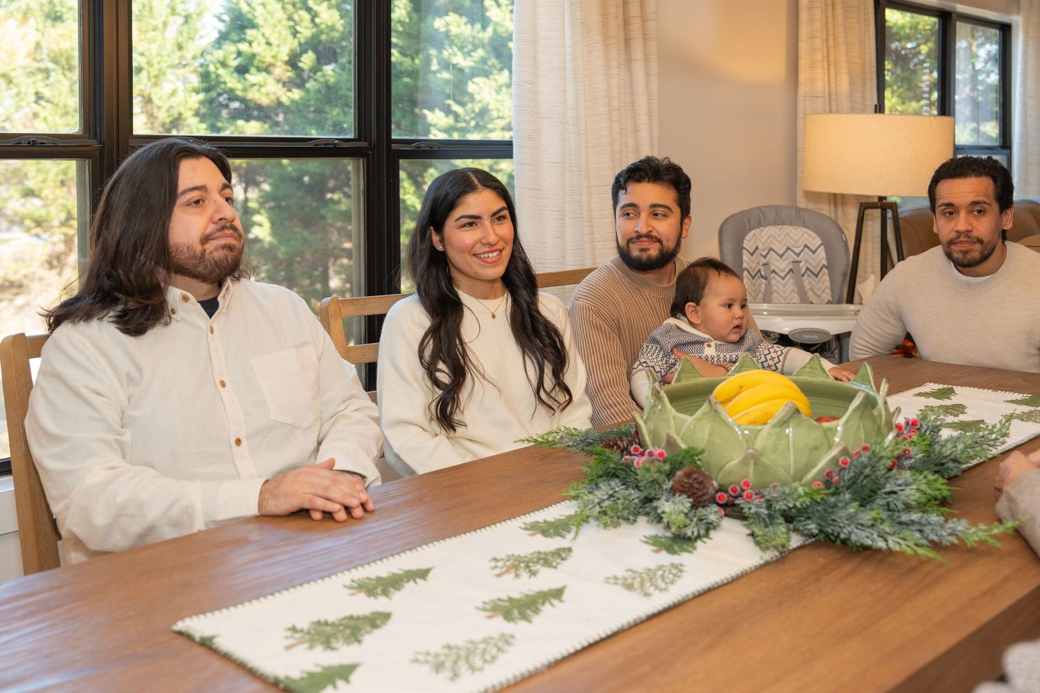 A group sits at a wooden dining table decorated with greenery during a gathering at home.
