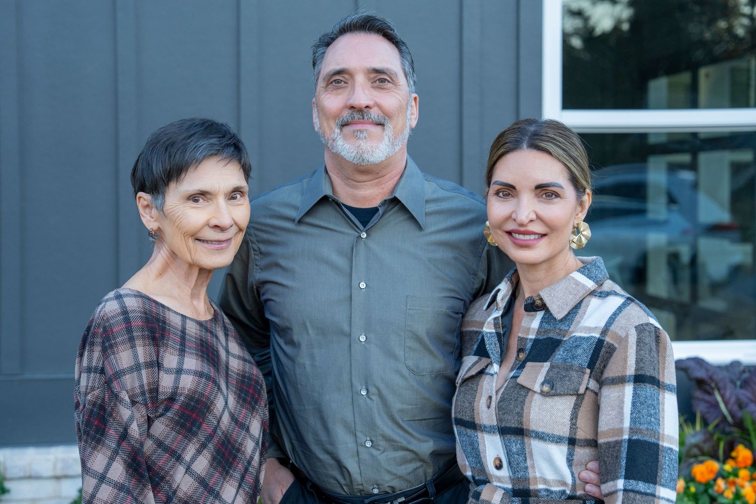 Three people stand smiling together outdoors against a blue building exterior with flowers visible in the background.