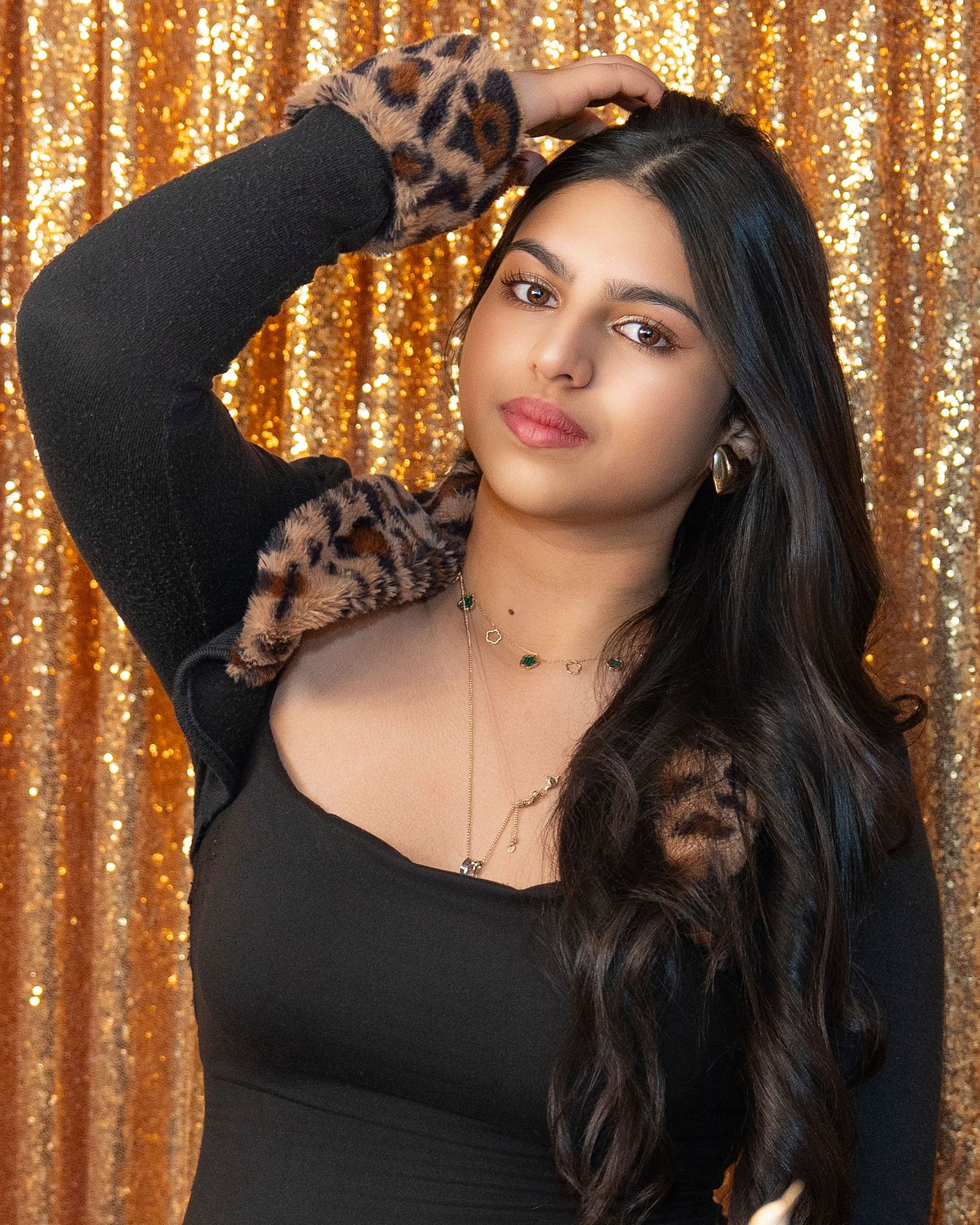 Woman in black long sleeve top poses against sparkly gold sequin backdrop.
