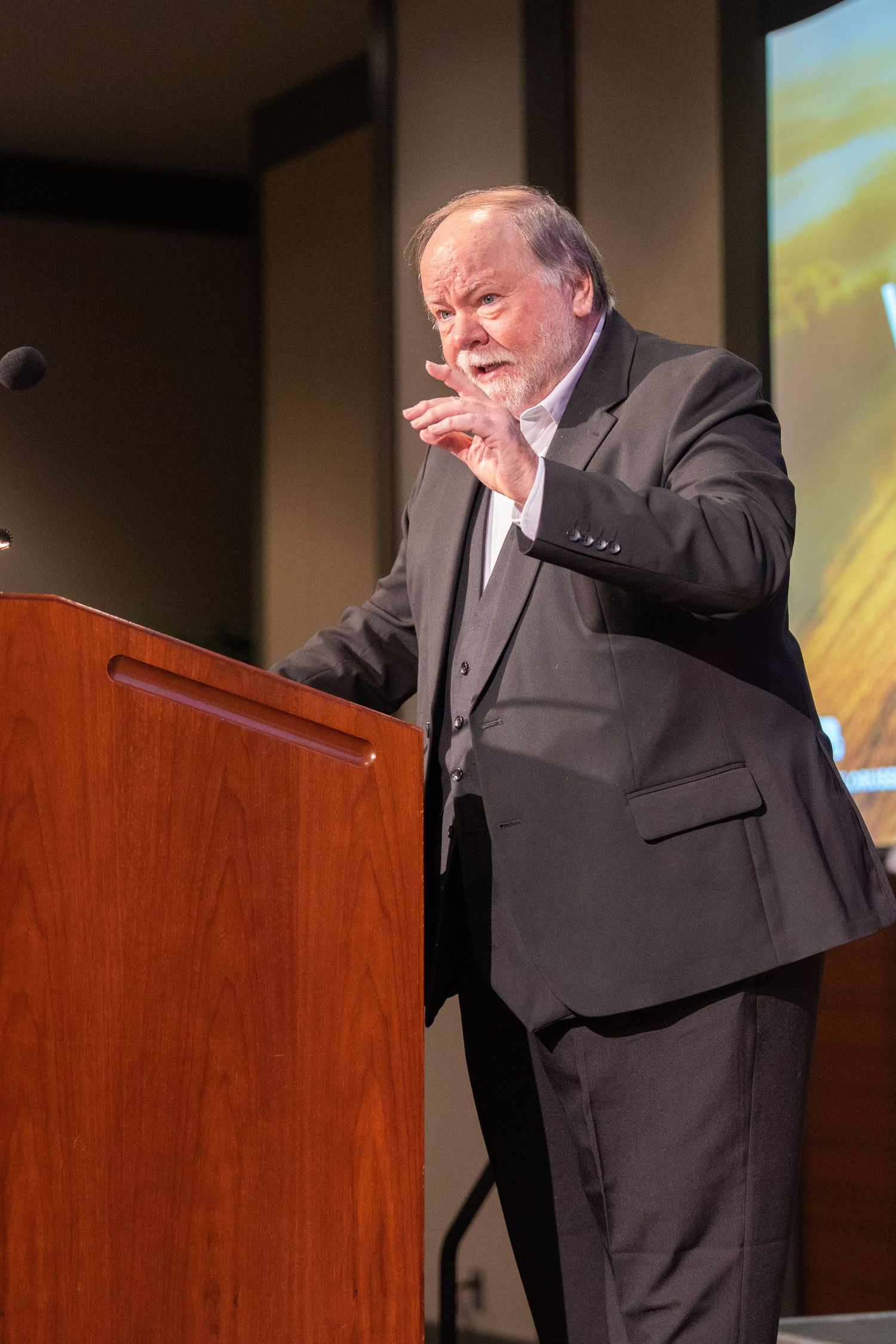 A speaker in a dark suit gestures while delivering remarks at a wooden podium during a presentation.