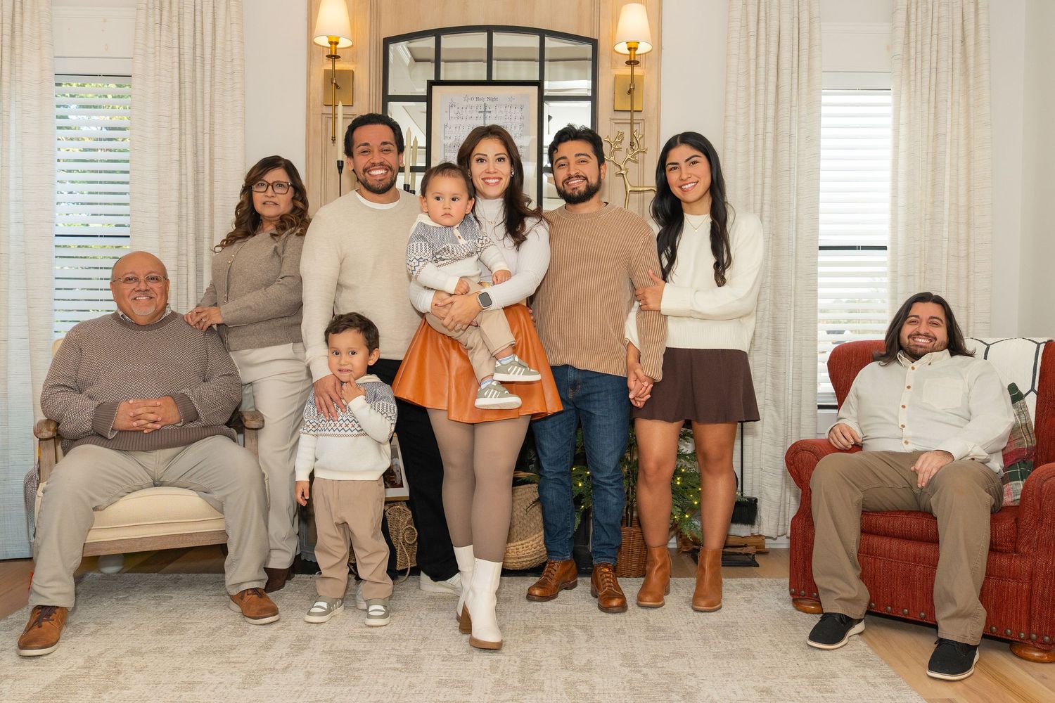 A large family group sits together on a beige couch in a bright living room with wall sconces and natural light.