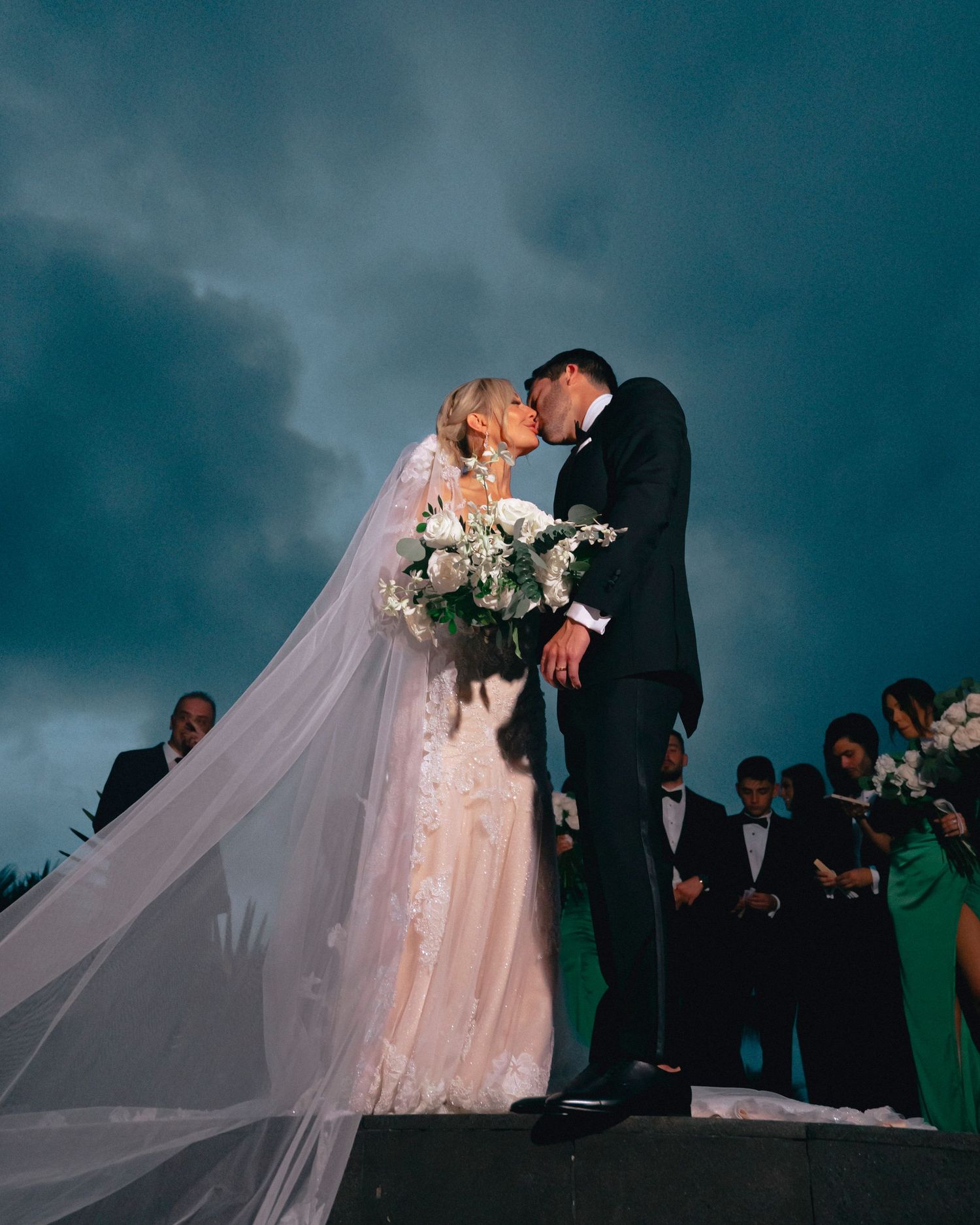 Romantic wedding couple embracing during sunset with dramatic teal sky backdrop.
