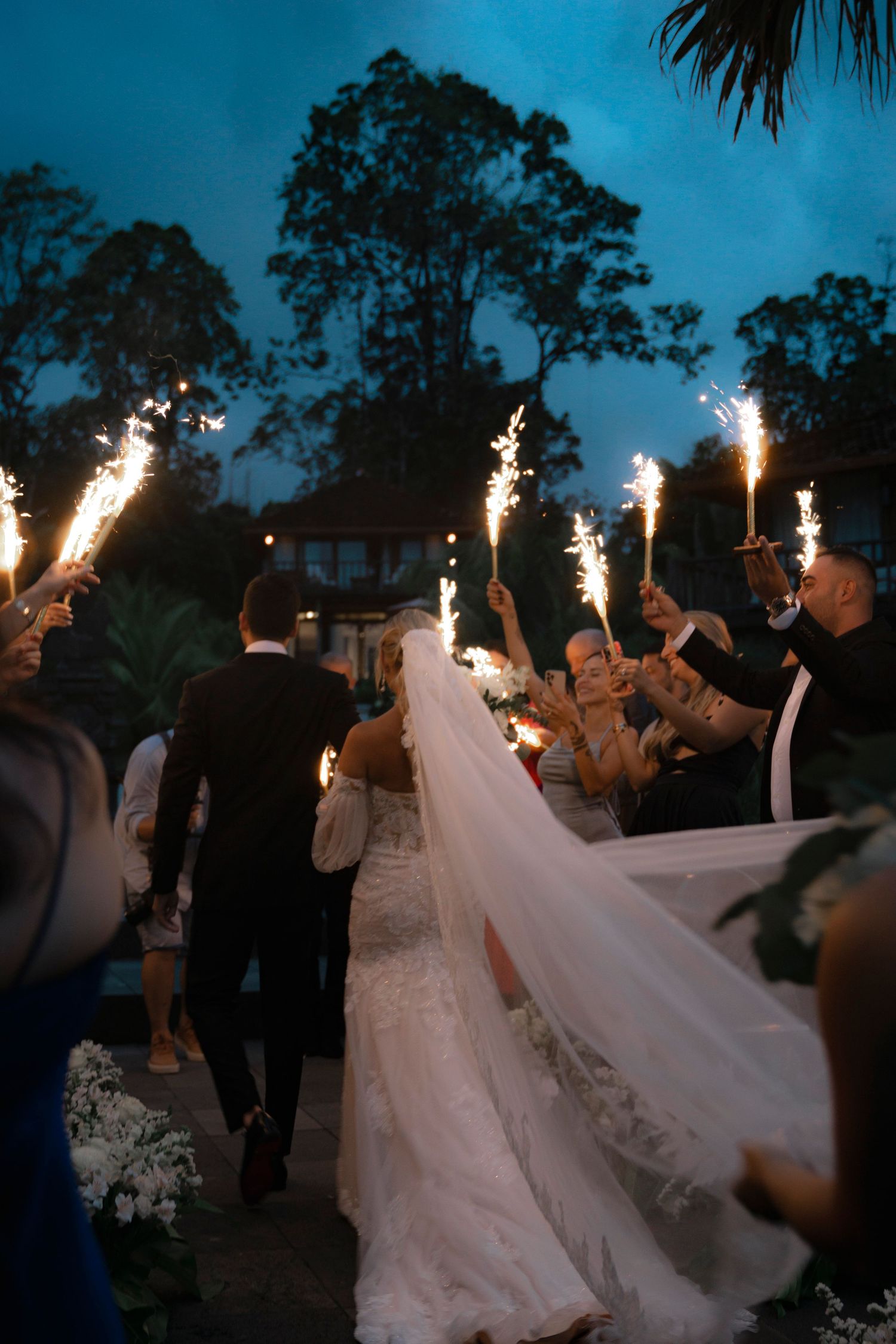 Wedding celebration at dusk with sparklers creating a magical exit tunnel for the newlyweds in flowing white dress.