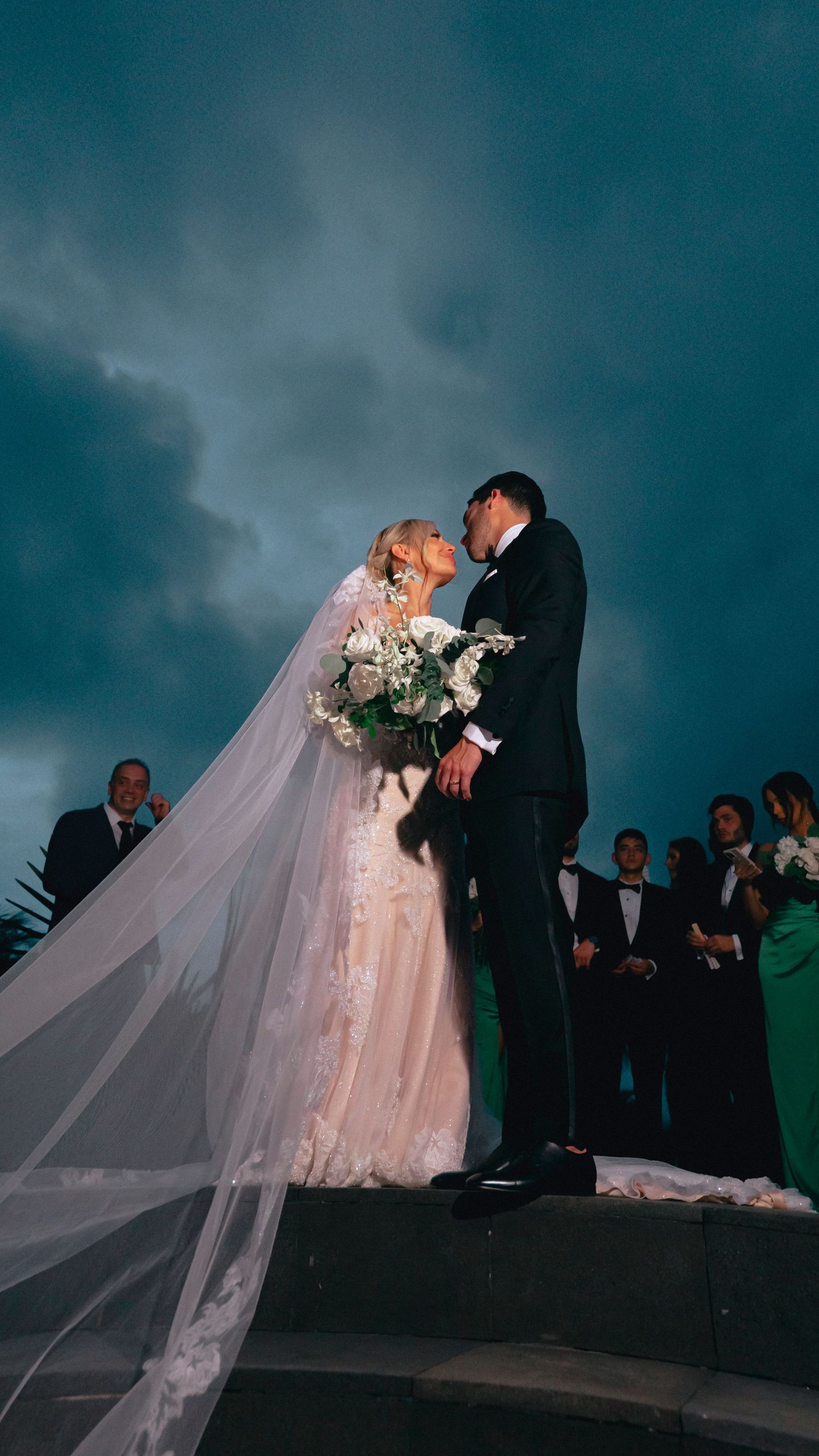Dramatic wedding kiss scene at dusk with flowing veil and guests in background.